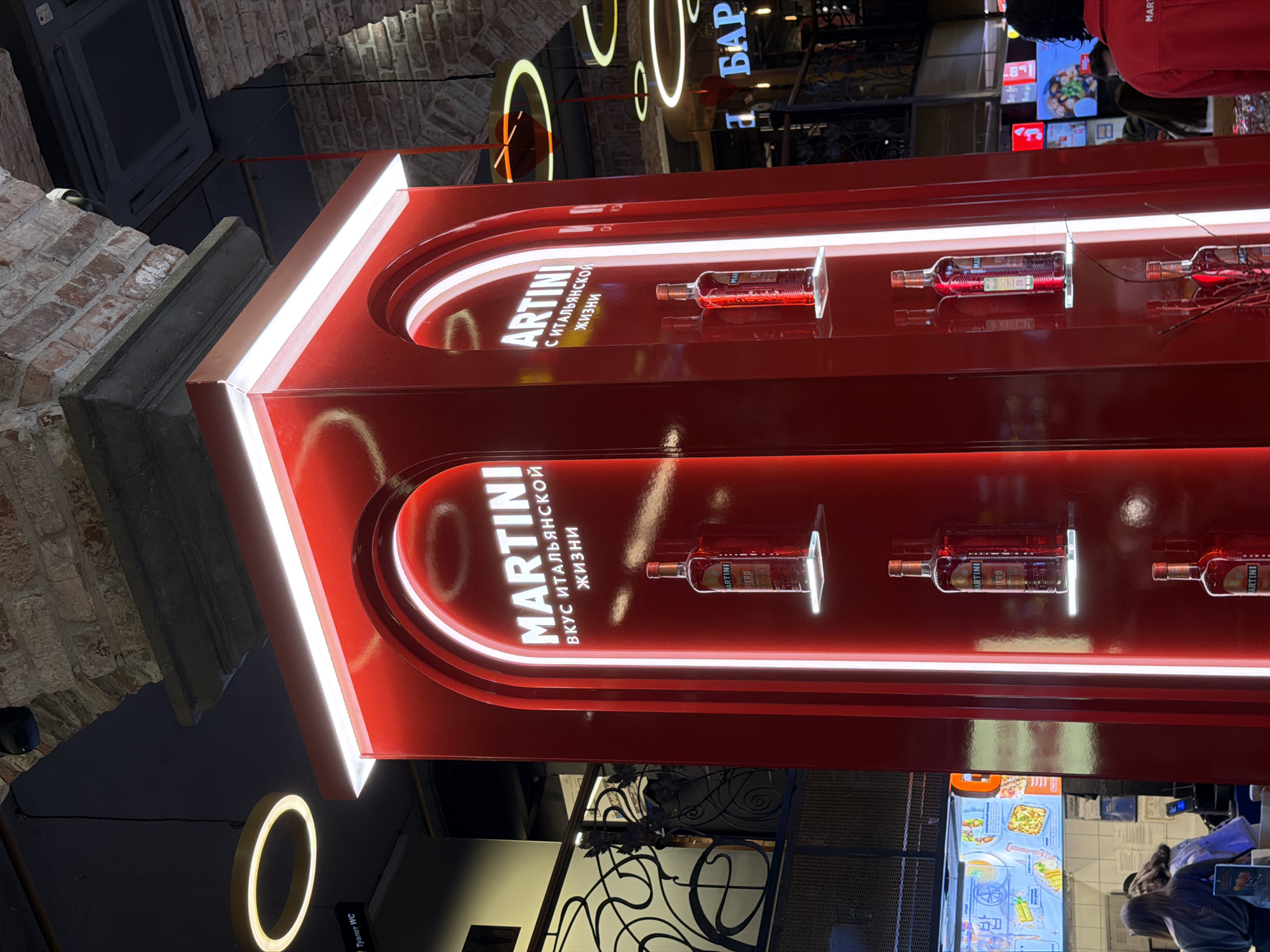 The image shows a bright red Martini display in what appears to be a public area, possibly a shopping center or food court. The display has two arched sections, each featuring bottles of Martini Fiero lit from behind. The text "MARTINI" is prominently displayed in Cyrillic letters, along with the phrase "Вкус итальянской жизни" (Taste of Italian life).
The background includes elements of the indoor environment, such as brickwork, lighting fixtures, and a glimpse of activity in the distance, including people and illuminated signs. The scene is busy, implying a crowded location.
Based on the Cyrillic script and the likely branding, the image is likely taken in Russia, possibly in Moscow.