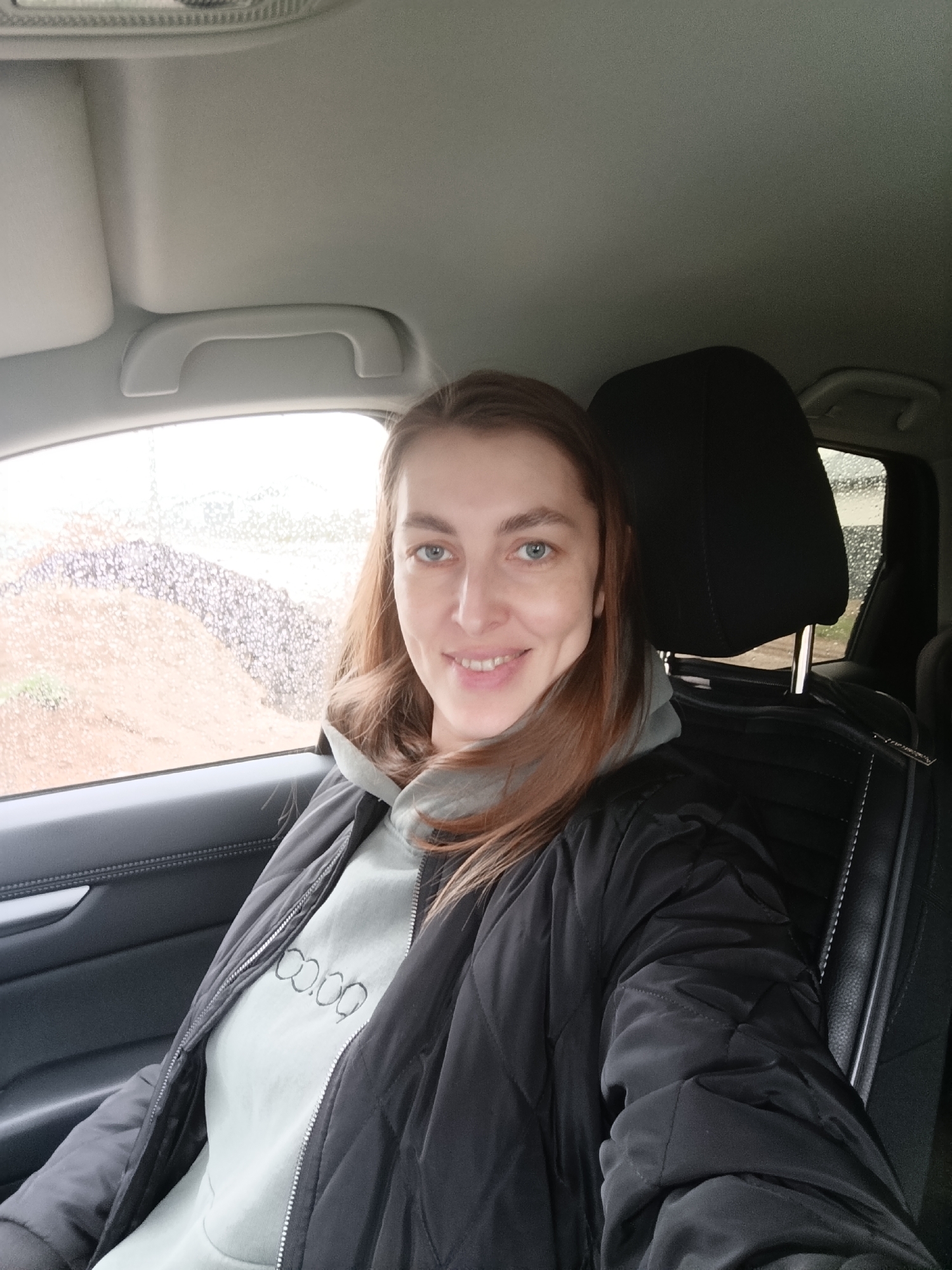 A woman is sitting in the driver's seat of a car. She is wearing a grey hoodie and a black quilted jacket. The window to her left is wet with rain. Outside the window appears to be a landscape of bare earth and vegetation, likely in a rural or undeveloped area. The interior of the car is visible, including the headrest of the driver's seat, the ceiling, and part of the dashboard. The photo appears to be a selfie taken by the woman. There is no definitive evidence that this image was taken in the city of Chaikovsky, Russia.