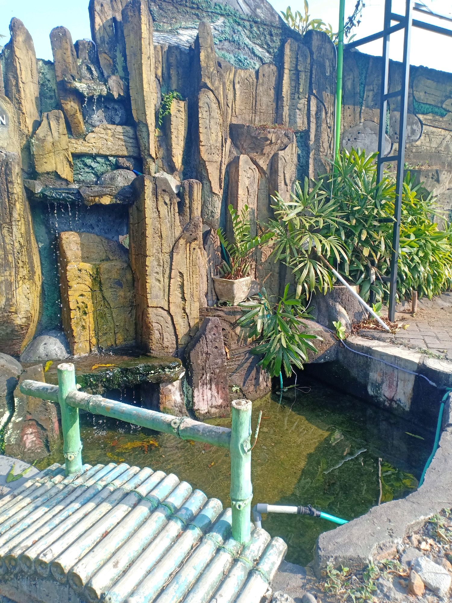 The image depicts a man-made rock waterfall feature with a small pond and a bamboo bridge in the foreground. The waterfall is constructed from textured material designed to resemble natural rock, with water cascading down in several places. The pond water is murky and reflects the surrounding environment. The bridge is made of light-colored bamboo poles, painted a pale blue and green, and is positioned over the pond. Several potted plants and lush green foliage are visible around the rock structure and behind it. A metal support structure is present on the right side of the image, along with a green hose running along the edge of the pond. The scene appears to be outdoors, with a bright sky suggesting daylight. The image was taken in the city of Pasirian, Indonesia, likely in a garden or park setting. There are no people visible in the image.