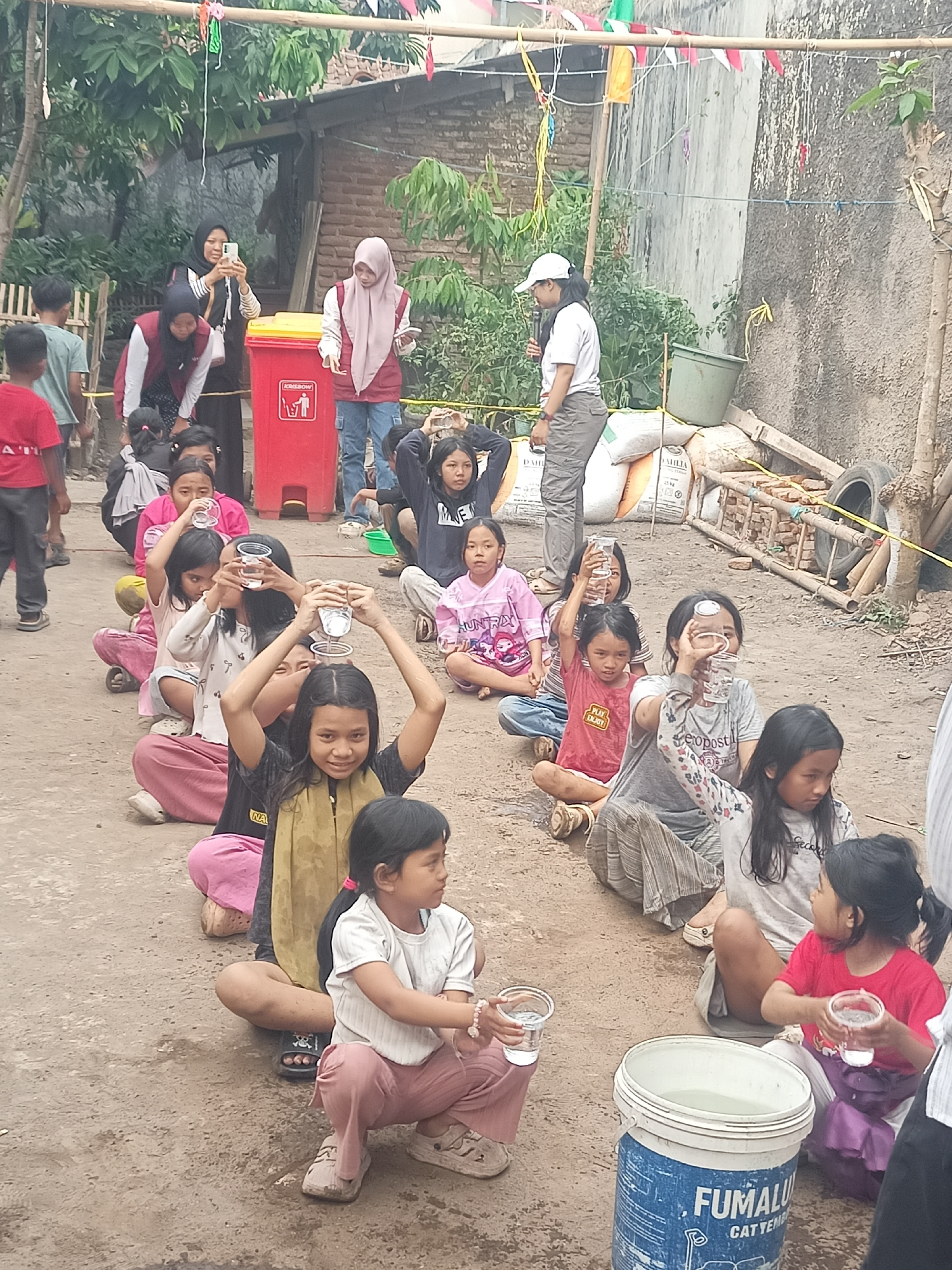 In a dirt yard in Tasikmalaya, Indonesia, a group of children are participating in an outdoor event. The children are mostly sitting in a line on the ground, with some holding plastic cups of water. One child in the foreground is wearing a yellow headscarf and a dark shirt, and is looking at the camera. Behind them, other children are also holding cups, some are raising them above their heads. In the background, adults are present, including a woman in a hijab taking a picture with her phone and another woman in a pink hijab holding a phone. There is a red trash bin, several bags of what appear to be cement or grain, and a large bucket filled with water. The scene appears to be an informal gathering or competition for children, possibly related to a community event or school activity, given the decorations of small flags strung overhead. The environment suggests a rural or semi-urban setting within Tasikmalaya.