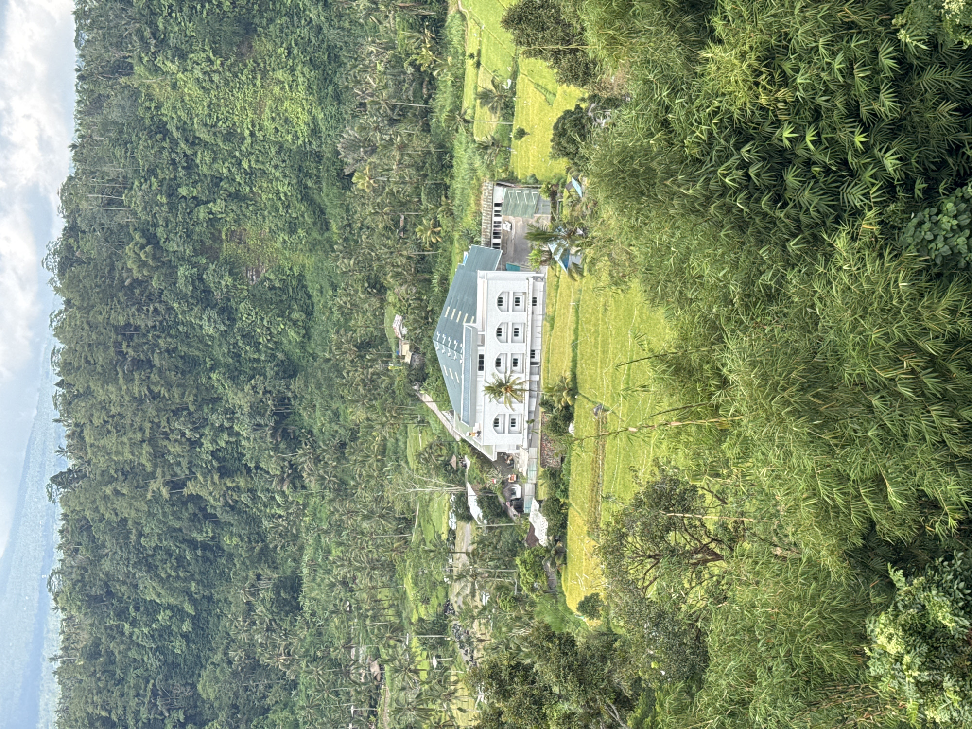 The image depicts a panoramic view of a landscape in Rendang, Indonesia. A multi-story, white building with a green roof stands prominently amid rice paddies and lush vegetation. The building has arched windows. A palm tree stands in front of the building. The area is surrounded by dense forests and hills, with glimpses of clouds in the sky. Smaller buildings are also present near the white building.