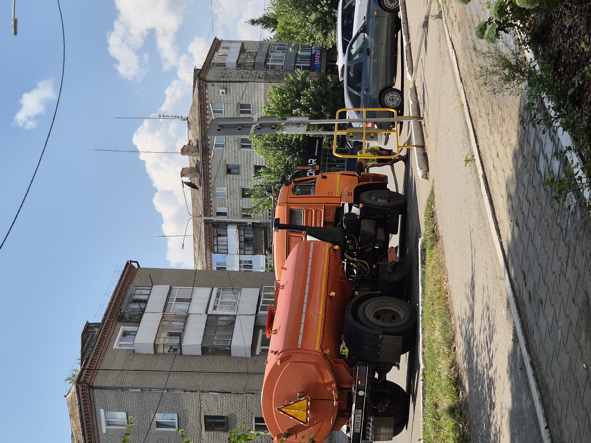 In Kokshetau, Kazakhstan, a bright orange water truck with a large cylindrical tank is parked on a paved road. The truck has the license plate "544 DA 01". A person wearing a bright yellow vest and dark shorts is standing near the truck, appearing to be involved in an operation with hoses connected to the truck. The truck is positioned in front of a multi-story brick apartment building with several balconies. Across the street, another apartment building is visible, as are several parked cars. The scene is under a bright blue sky with scattered white clouds. Trees and greenery are present on the sides of the road.