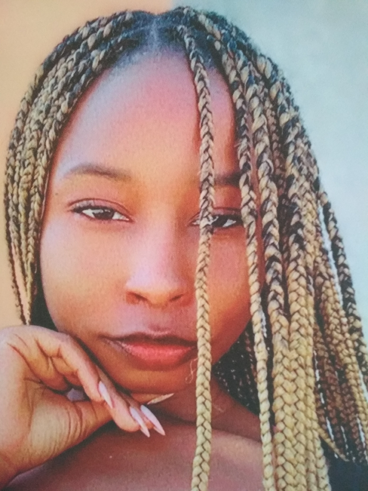 A close-up portrait features a woman with brown skin. She is looking directly at the viewer with a slightly contemplative expression. Her hair is styled in long, braided extensions that are a blend of blonde and black. A strand of hair falls directly in front of her face, partially obscuring one eye. Her left hand is positioned near her chin, with long, manicured nails visible. The background is a blurred gradient of warm, neutral tones. No information about a location in the city of Unknown, Unknown can be discerned from the image.