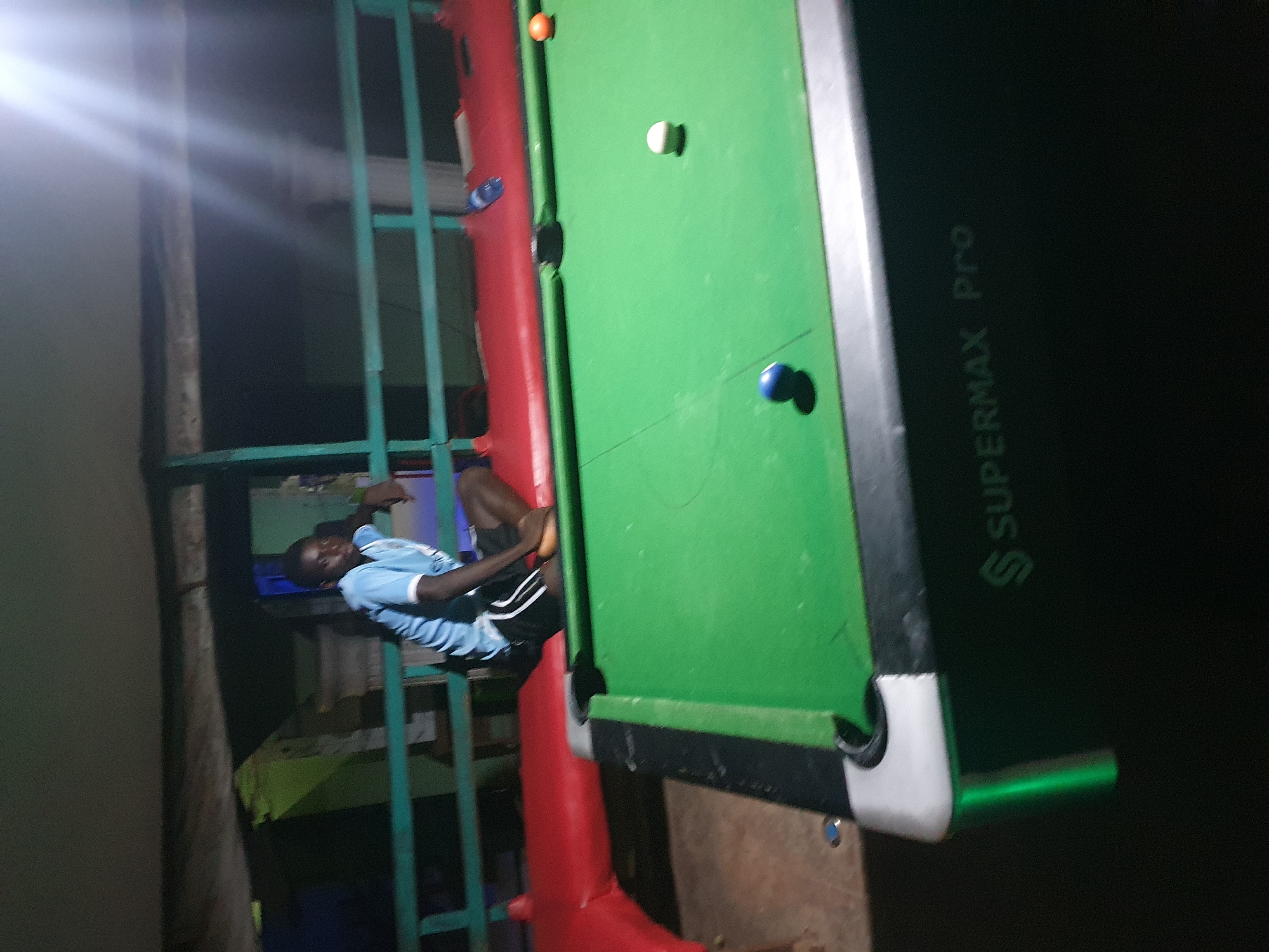 A young Black male wearing a blue and white t-shirt and black shorts is sitting on a red inflatable border that surrounds a green pool table. He is looking towards the left of the frame. The pool table has three visible balls: a white ball, a blue ball, and an orange ball. The pool table is labeled "SUPERMAX Pro". The background appears to be a dimly lit, outdoor or semi-outdoor area with green railings and some structures. The scene suggests a recreational setting, possibly a pool hall or a backyard game area.