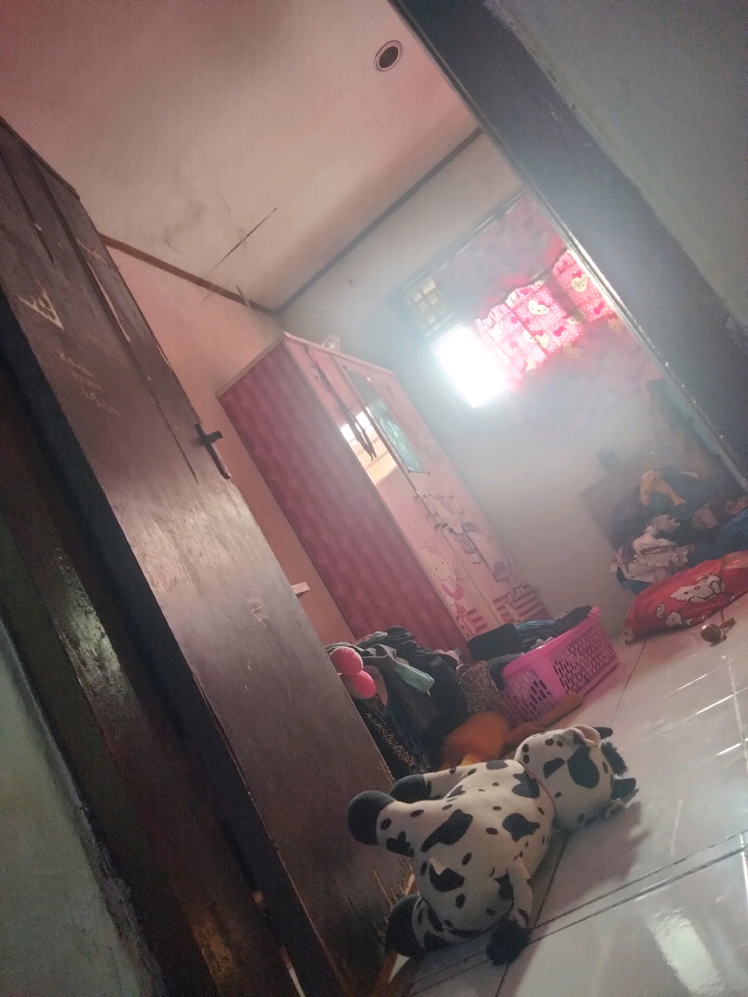 The image depicts a room interior, possibly a bedroom, with a tilted perspective. A wooden door dominates the left side of the frame, appearing old and worn. In the background, a pink wardrobe with a cartoonish design is visible. To the right, a window is covered by a pink curtain featuring a pattern of Hello Kitty characters. Various items are scattered around the room, including clothes, a pink basket, and a plush toy resembling a cow lying on the tiled floor. The lighting suggests it is daytime, with sunlight streaming through the window. The overall impression is of a somewhat cluttered and lived-in space. The location is specified as Suka Sari, Indonesia.