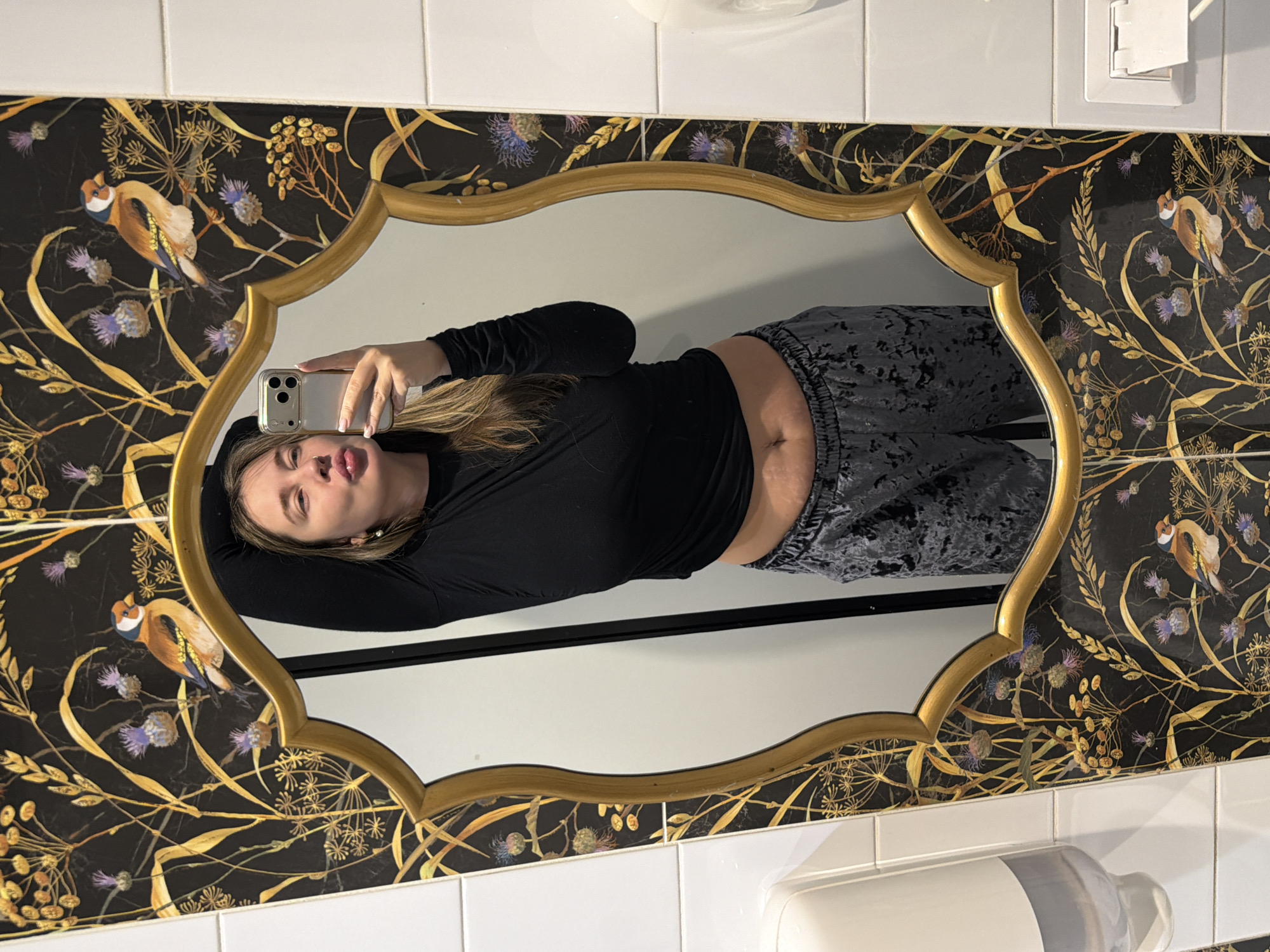 A woman is captured in a mirror selfie, with the image rotated 90 degrees counter-clockwise. She lies horizontally within the gold-framed mirror, holding a gold-colored smartphone to take her picture. Her long, light brown hair cascades around her shoulders. She is dressed in a black long-sleeved top that reveals a sliver of her midriff and dark grey or black patterned loungewear or sweatpants. Her expression is neutral, with slightly parted lips.
The mirror itself has a distinctive ornate gold frame with a scalloped edge. Surrounding the mirror is an eye-catching black wallpaper adorned with intricate gold and yellow botanical designs, including delicate foliage, seed heads, and various purple thistle-like flowers. Several small birds, resembling goldfinches, are depicted perched among the plants on the wallpaper.
The setting appears to be a well-decorated bathroom or powder room, indicated by the white square tiles visible above and below the wallpaper, and a white, wall-mounted soap dispenser or similar fixture partially seen in the bottom right corner. A white electrical outlet or switch cover is also visible in the top right. The overall aesthetic suggests a stylish interior, located in Moscow, Russia. No discernible text is visible in the image.