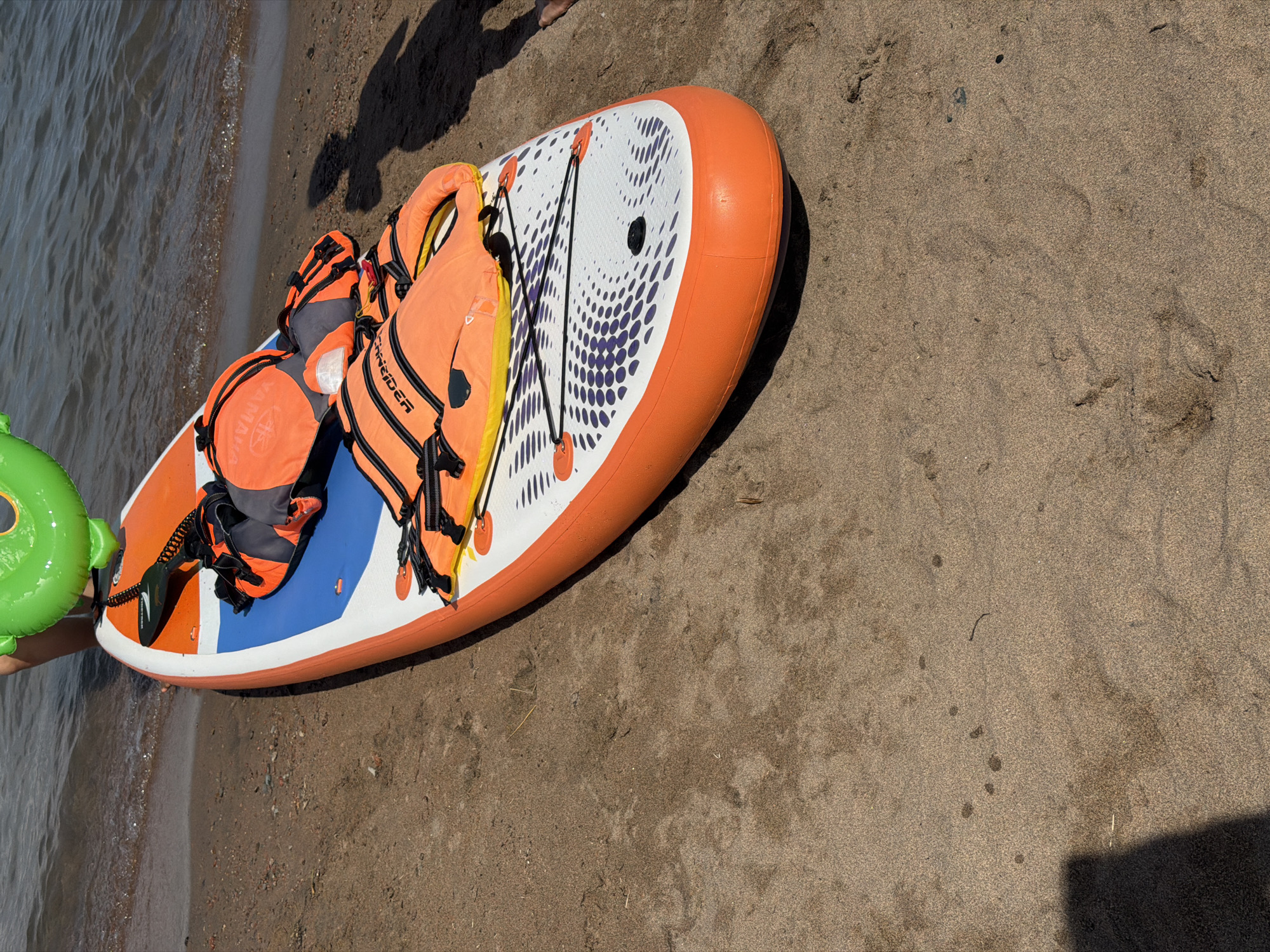 An orange and white inflatable stand-up paddleboard rests on a sandy beach next to the water. Two orange life vests are placed on top of the paddleboard. A person's legs and a portion of a green inflatable ring are visible in the upper left corner of the image, near the water. The scene is set at a beach on Issyk-Kul Lake in Choktal, Kyrgyzstan.