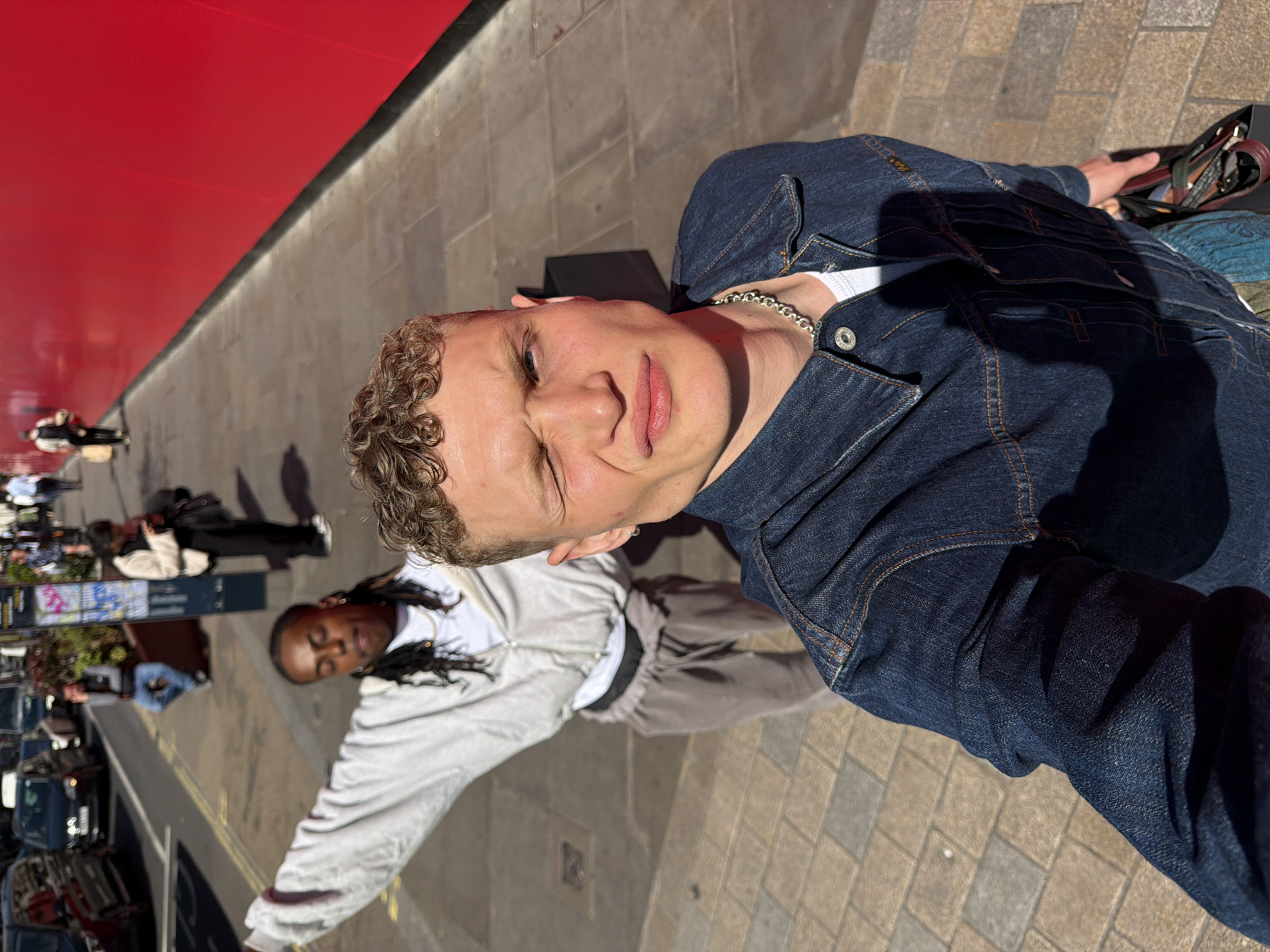A young man takes a selfie on a bright, sunny day in what appears to be a city street in London, United Kingdom. He is a white male with light brown curly hair, looking directly into the camera with a slight squint, likely due to the sun. He wears a dark blue denim jacket with orange stitching over a white t-shirt and a silver chain necklace. His lips are slightly parted, and his expression is relaxed. A black strap or bag is visible near his right arm.
Behind him, slightly out of focus, stands another person with dark skin and dark hair, possibly styled in braids or dreadlocks. This individual is wearing a light grey zippered jacket and light grey sweatpants, looking downwards or away from the camera.
The setting features light-colored block paving underfoot. A striking, vibrant red wall or building façade occupies the left side of the frame, creating a strong diagonal line. Prominent shadows are cast across the ground and subjects, indicating strong sunlight, consistent with daytime. In the blurred background, other pedestrians, parked cars, and indistinct street furniture or signs suggest a busy urban environment. No legible text is visible.