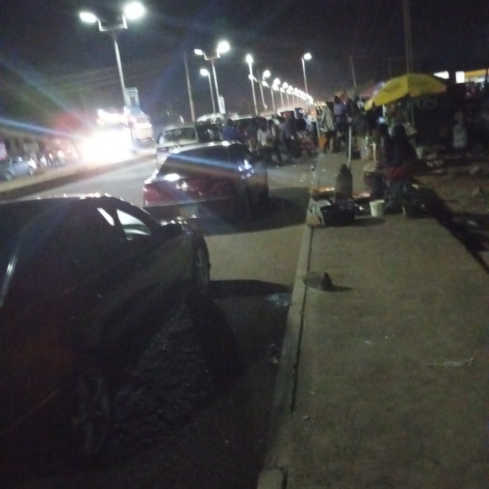 At night in Bukuru, Nigeria, a street scene reveals a row of illuminated streetlights lining a road with parked cars and pedestrians. A yellow awning covers a roadside stall where people are gathered. On the pavement, cooking utensils and containers sit near the edge. Dark cars are parked on the road shoulder, with a stream of headlights visible in the distance.