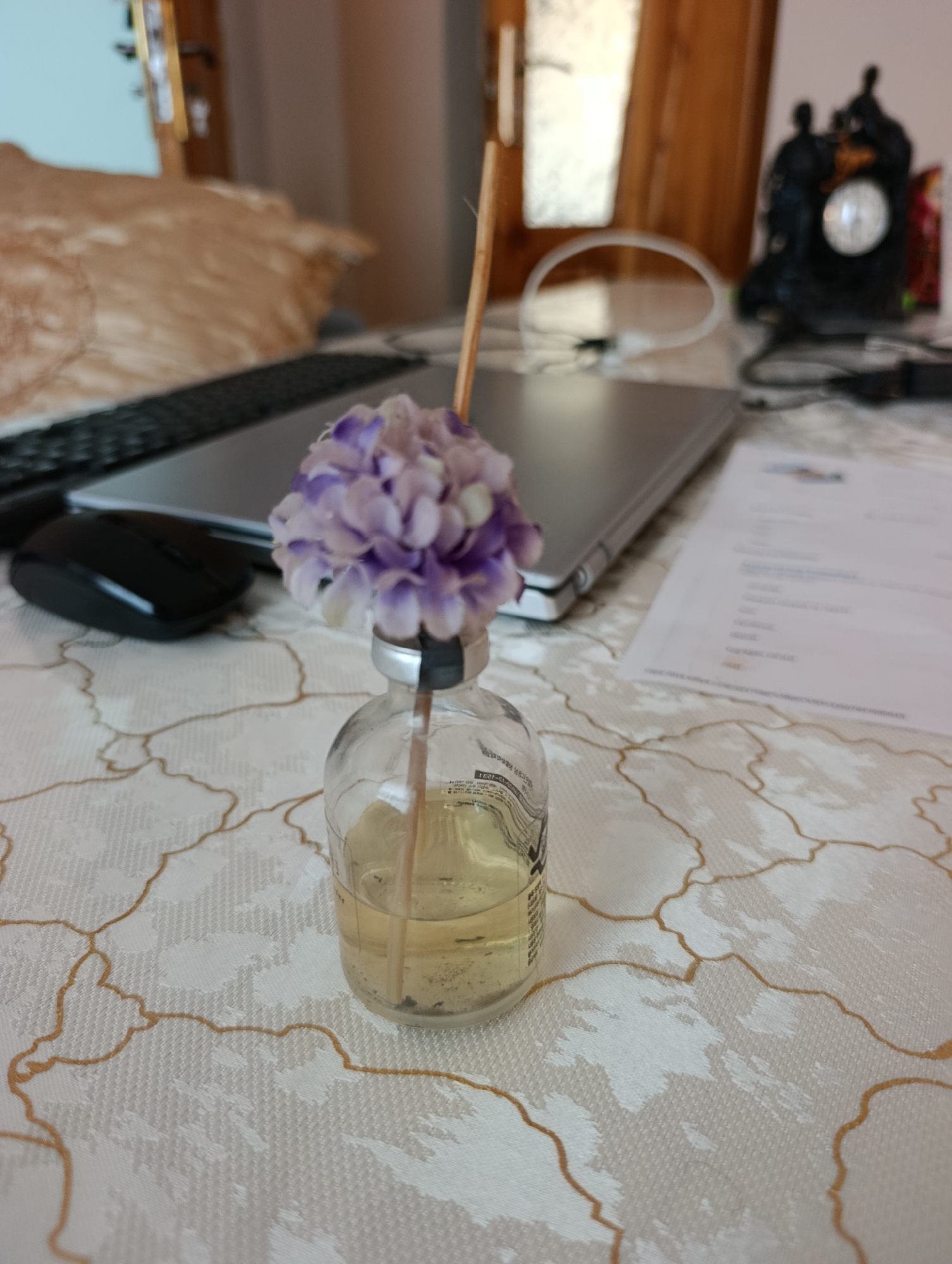 The image displays a close-up of a small glass bottle containing a pale yellow liquid and a single reed diffuser stick. A cluster of artificial purple and white flowers is attached to the top of the reed. The bottle rests on a white tablecloth with an intricate gold pattern. In the background, a silver laptop, a black computer mouse, and a blurred antique clock are visible. The scene appears to be an indoor setting, possibly a desk or table, but there is no information to suggest its location in Laish, Uzbekistan.