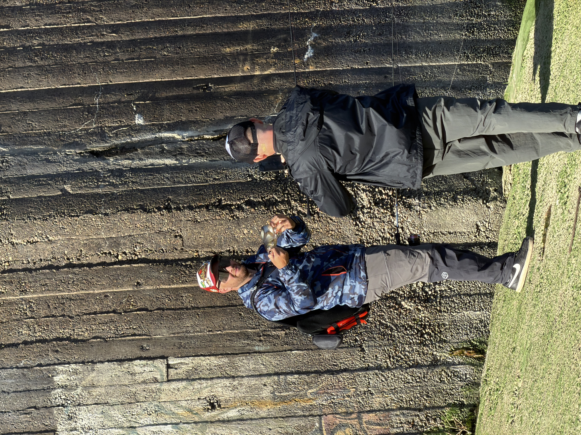 Two men are positioned adjacent to a textured, concrete retaining wall. The wall exhibits horizontal lines or seams. One man, wearing a blue camouflage jacket and red-trimmed cap, is holding a fish. He is also wearing sunglasses, grey pants, and has a bag beside him. The other man is dressed in a black jacket and grey pants, and is wearing a black and white baseball cap. The ground surface consists of small gravel and some grass. This is presumed to be in Chaikovsky, Russia.