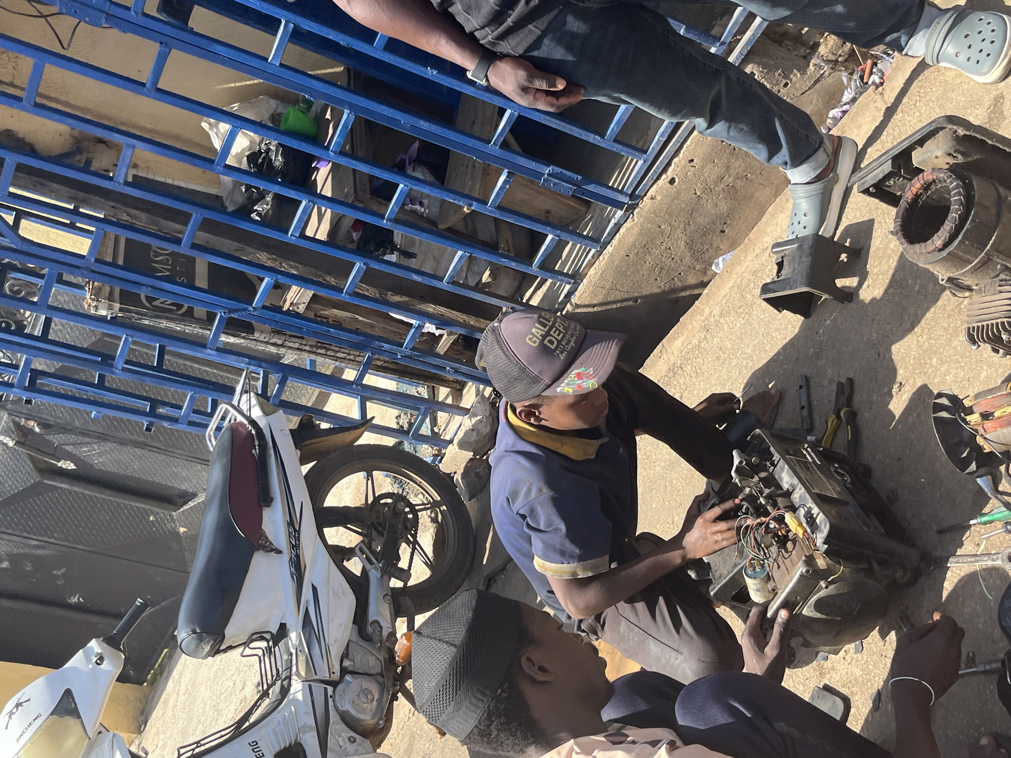 In the city of Jos, Nigeria, a group of individuals are working on a generator. The scene appears to be outdoors, possibly in a workshop or a roadside repair area, with a white motorcycle parked to the left and a blue metal fence to the right. The ground is a rough, concrete surface.
One person, wearing a blue shirt and a dark baseball cap, is crouched down, actively engaged with the generator's internal components. Their hands are manipulating wires and parts of the machine. Another individual, wearing a light-colored shirt and a black cap, is also looking at the generator, with their hands near the machine. A third person, visible from the waist down and wearing blue jeans and grey Crocs, stands to the right, observing the work.
The generator itself is a black, rectangular unit with visible wiring, coils, and metal parts. Tools such as pliers and screwdrivers are scattered around the generator on the ground, indicating active repair work. Parts of other dismantled machines are also present in the foreground. The overall atmosphere suggests a practical, hands-on repair or maintenance operation.