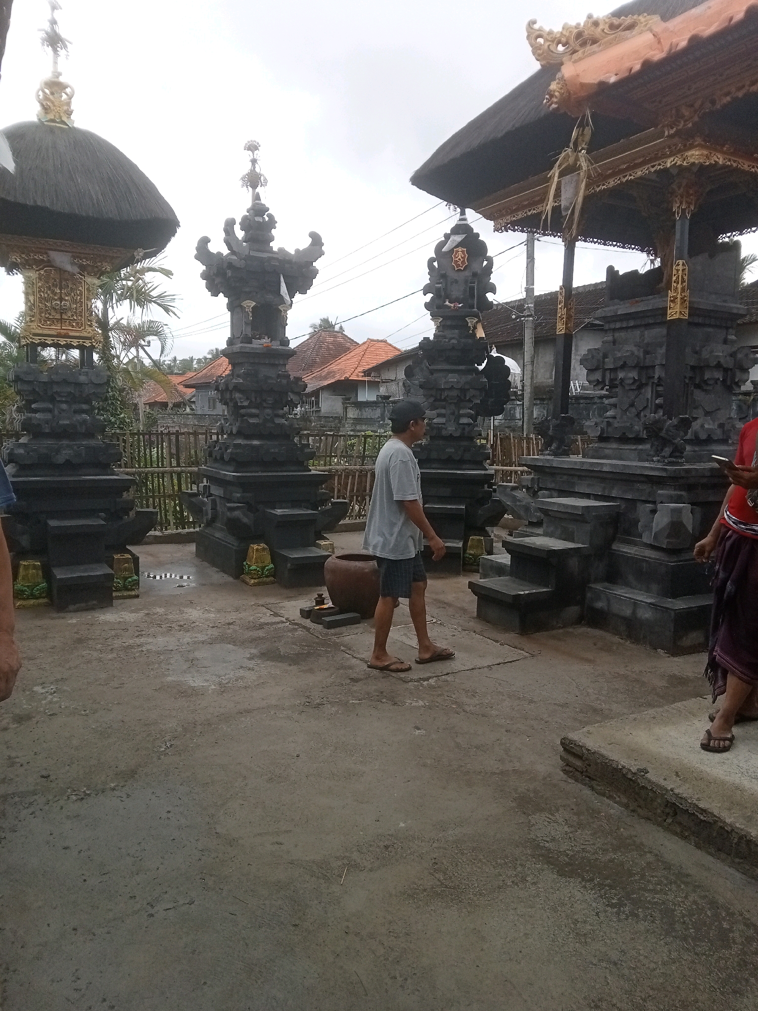 In Mendoyo, Indonesia, at what appears to be a Balinese temple complex, a young man in a grey t-shirt and shorts walks across a concrete courtyard. He is heading towards the right side of the frame, away from the viewer. To his left are two intricately carved stone structures, likely part of a temple gate or shrine, topped with dark thatched roofs and gold accents. A large, dark brown clay pot sits on the ground near the stone structures. To the right, a more elaborate structure with a dark thatched roof and gold decorative elements is visible, with stone steps leading up to it. Another person, wearing a dark skirt and a red top, stands to the far right, looking down at their phone. The background consists of residential buildings with red tile roofs and palm trees, suggesting a rural or semi-urban setting. The sky is overcast, indicating a cloudy day.