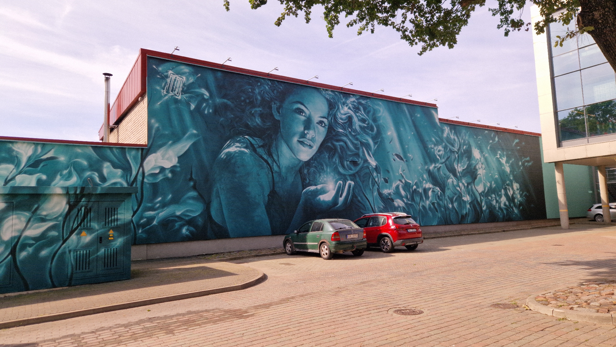 The image depicts a large mural on the side of a building in Liepaja, Latvia. The mural features a woman's face with flowing hair, surrounded by aquatic elements like fish and plants, rendered in shades of blue and green. In front of the mural, two cars are parked: a green hatchback and a red SUV. The building itself has a red-trimmed roof and a brick section. To the right, a modern building with large glass windows is visible. The foreground is a paved area with cobblestones and a sidewalk. The scene is bathed in daylight, casting shadows on the ground.
