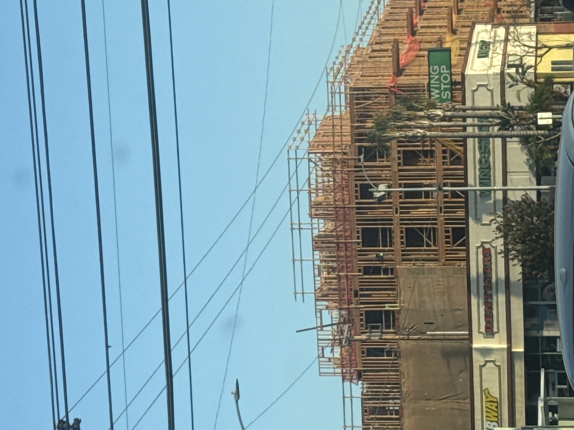 The image shows a construction site in Los Angeles, California, United States of America. The scene is dominated by a multi-story building under construction, with its wooden frame exposed. Various scaffolding and safety nets are visible. Below the construction, a row of businesses appears to be in operation. Signs for "SUBWAY" and "DENTISTRY" are clearly visible. A green sign with "WING STOP" is also present, suggesting a fast-food restaurant. Several palm trees are scattered around the area, a common sight in Los Angeles. Overhead, power lines crisscross the clear blue sky, and a street light is also visible. The overall impression is of urban development in progress.