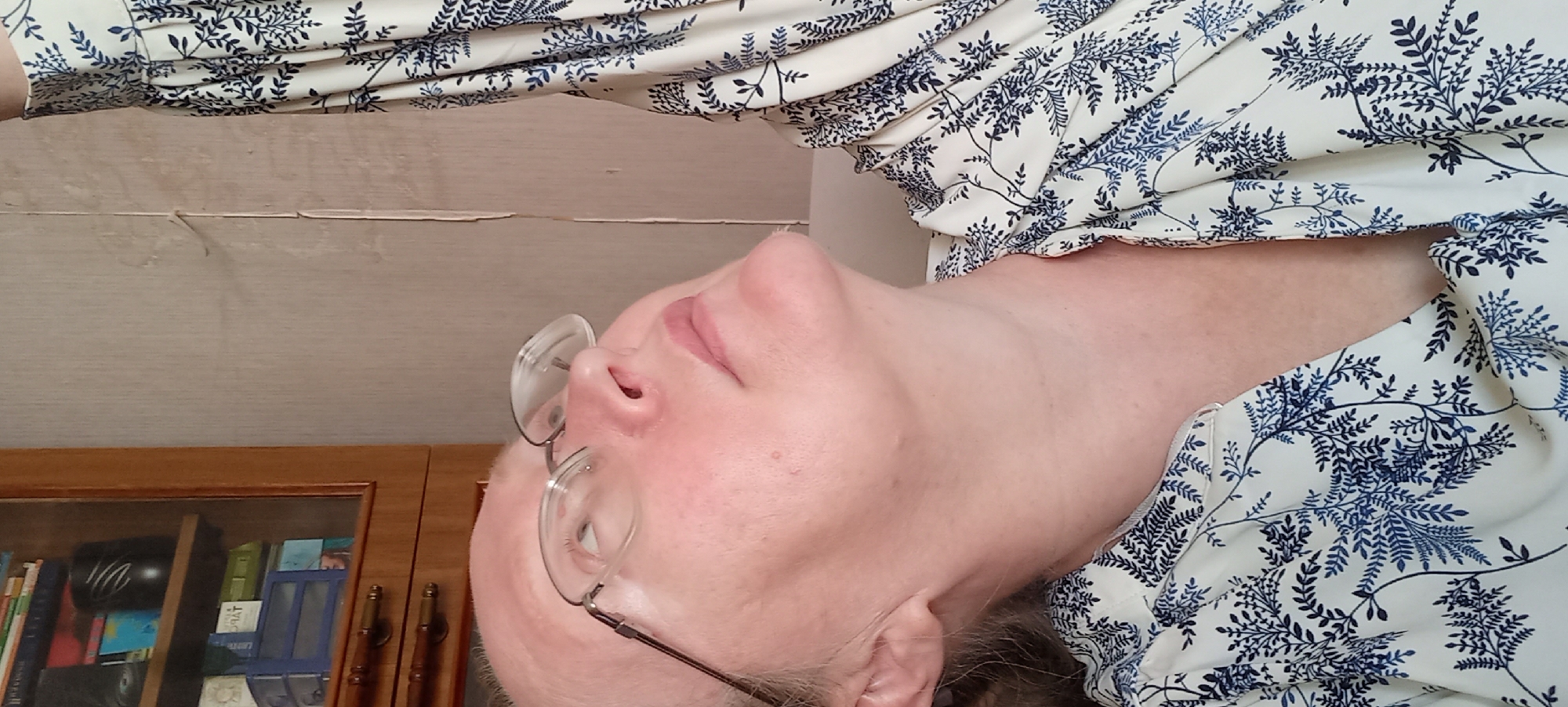 The image is a close-up, vertically oriented shot of a person's face and upper body. The subject is wearing glasses and a cream-colored shirt with a navy blue leafy pattern. The person's head is tilted back, with their face looking upwards. The lighting suggests an indoor setting, likely during the day. The background includes a wooden shelf with several books and decorative items, and a textured wall above it. The overall impression is a candid, possibly self-taken photograph. There is no visible text in the image.