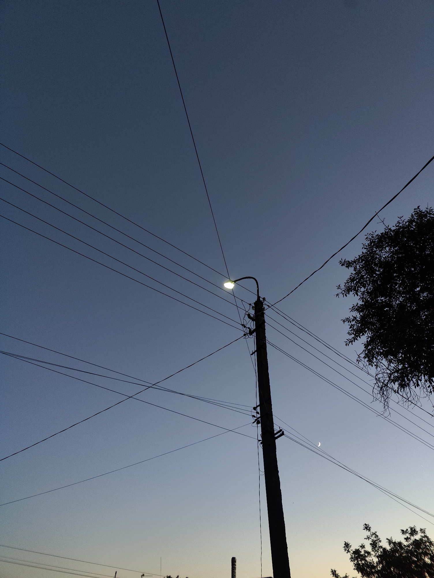 A power pole with a street light and multiple wires stands silhouetted against a twilight sky in Yarmolyntsi, Ukraine. A crescent moon is visible in the sky. A tree branch is also silhouetted against the sky.