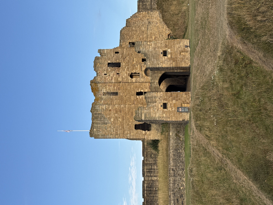A ruined stone castle, located in North Tyneside, United Kingdom, stands under a clear blue sky. The structure is composed of light brown, cut stone blocks and displays significant architectural wear and decay, with a prominent central keep and connecting curtain walls. Multiple rectangular and arched openings are visible throughout the weathered facade, some appearing as empty window apertures and others as barred recesses. A large, dark arched gateway serves as the primary entrance, piercing the central lower portion of the structure. A white flagpole, topped with a partially furled or indistinct flag displaying red, white, and blue, consistent with the Union Jack, extends from the upper left battlements. The immediate foreground consists of uneven, dry grassy terrain, marked by two visible dirt paths ascending towards the castle's base. To the left of the main archway, a dark information sign is affixed to the stone wall. The scene is devoid of human or animal presence. The lighting is natural, suggesting daytime.