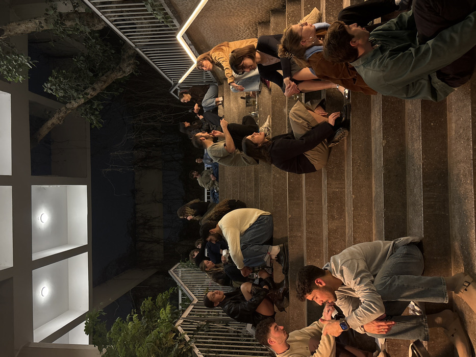 A group of people sits on outdoor stairs, stretching out across multiple steps. Some individuals are paired and appear to be engaged in conversation or looking at phones. The scene is lit dimly by artificial lights from a building and railing. A tree is to the left of the stairs. A dark sky is visible in the background.