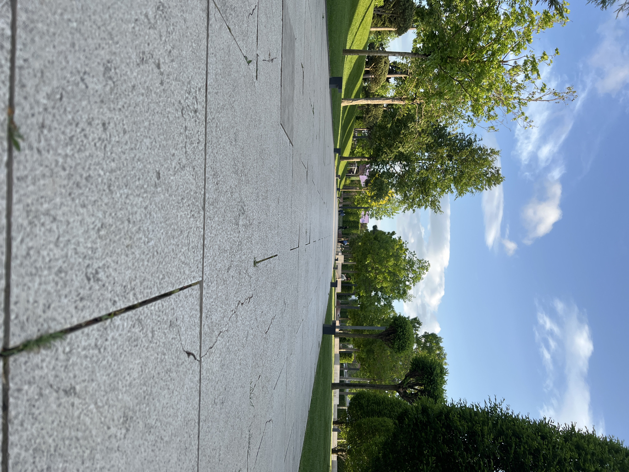 A pathway made of large, light gray rectangular stones extends into the distance, framed by green grass on the right and trees on the right. The sky is partially visible, with white clouds and patches of blue. The image captures an outdoor public space with an architectural path and curated greenery. Given the location in Krasnodar, Russia, it is likely a park or memorial area with landscaped design. No individuals are clearly visible.