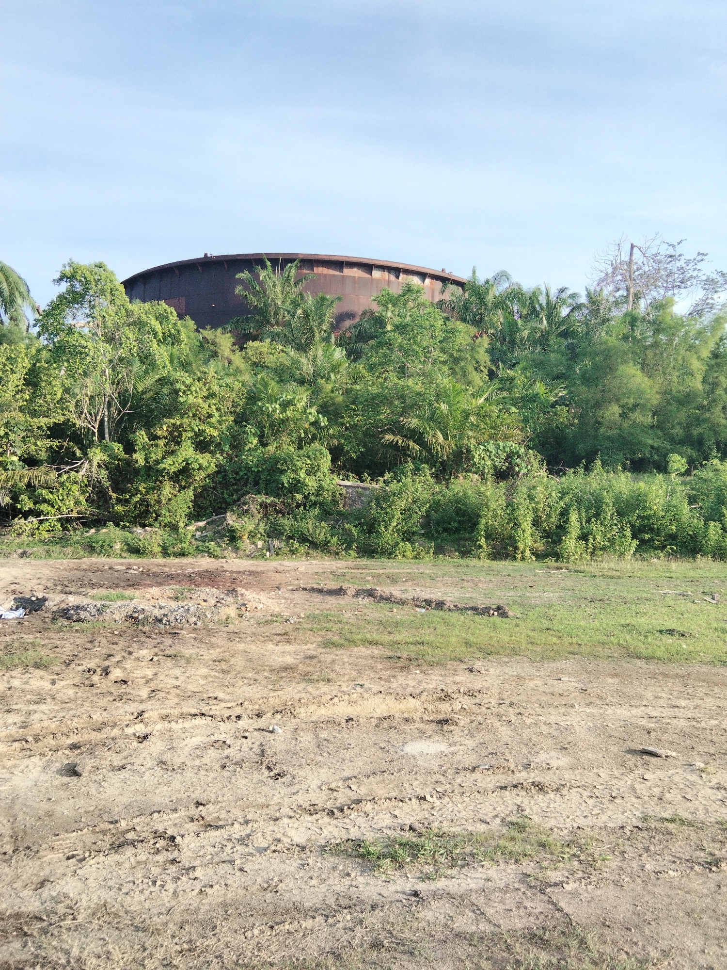 The image depicts an outdoor scene under a clear blue sky. In the background, a large, circular, rusted metal structure, possibly a water tower or industrial storage tank, is partially visible above a dense canopy of lush green trees and vegetation. The foreground consists of a patch of dry, uneven ground with tire tracks and scattered debris, transitioning into a grassy area. The overall impression is of an undeveloped or abandoned industrial site encroached upon by nature. The location is specified as Sukaramai Dua, Indonesia, suggesting a tropical or subtropical environment given the dense foliage. There are no people or animals visible in the frame.