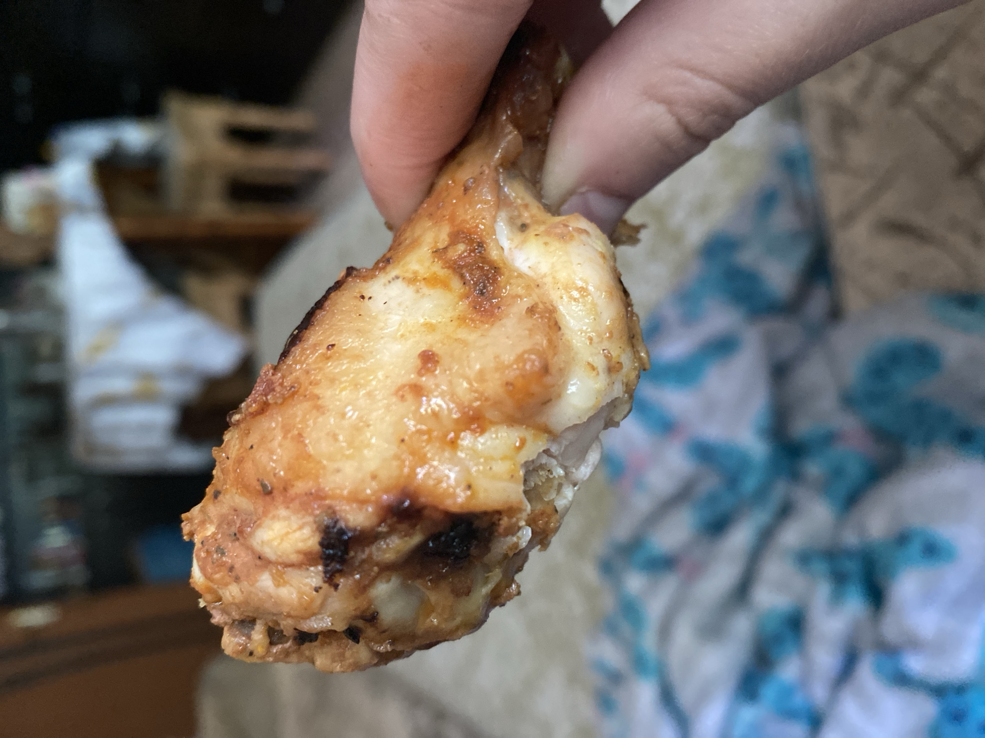 A close-up shot shows a hand holding a piece of cooked chicken wing. The chicken wing is golden brown with some dark, slightly charred spots. The skin appears crispy, and some of the white meat is visible. The background is blurred and out of focus, but it appears to be an indoor setting with what looks like furniture and possibly bedding. The lighting suggests it is daytime.