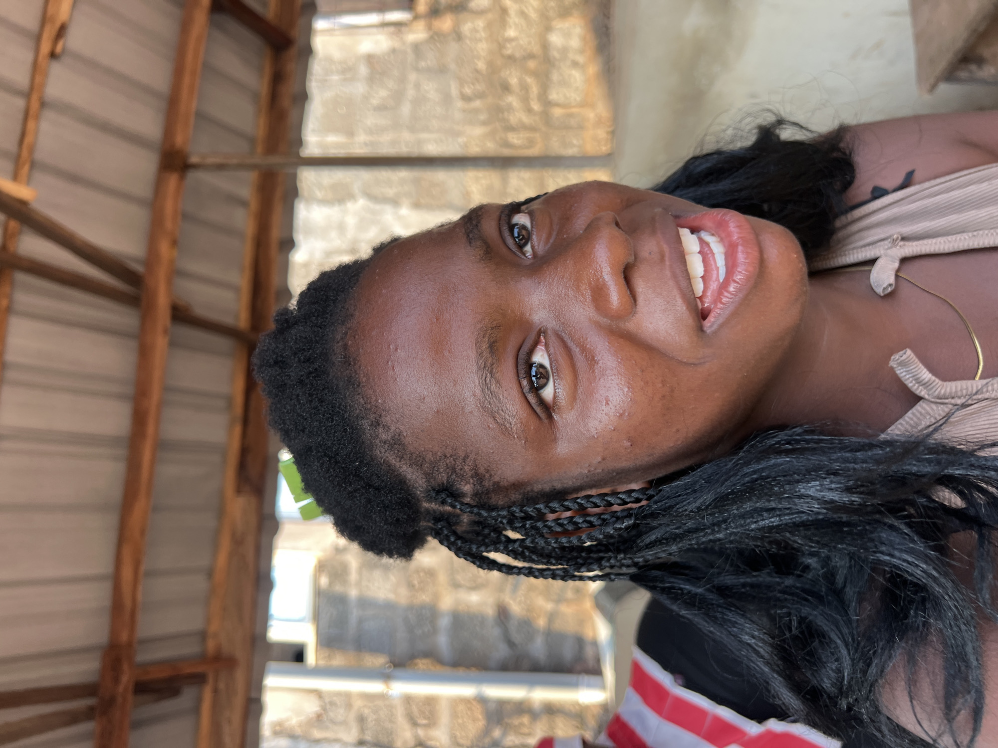 A medium close-up shot features a young Black woman with dark skin, dark eyes, and black braided hair, a few strands of which are loose and fall around her shoulders. She is looking slightly to her right, with a wide smile that reveals her teeth and pink gums. Her lips are glossed and appear full. She is wearing a light beige, ribbed tank top with thin straps. A thin gold necklace is visible around her neck. Her skin has a slight sheen, suggesting it may be damp. A green leaf is tucked into her hair near the top of her head. Behind her, a structure with a corrugated metal roof supported by wooden beams is visible. The background shows a rough, light-colored stone wall and a white pipe. To her left, a person wearing a red and white striped vest over a black shirt is partially visible. The scene appears to be outdoors, possibly in a market or workshop, consistent with the urban environment of Nkpor, Nigeria.