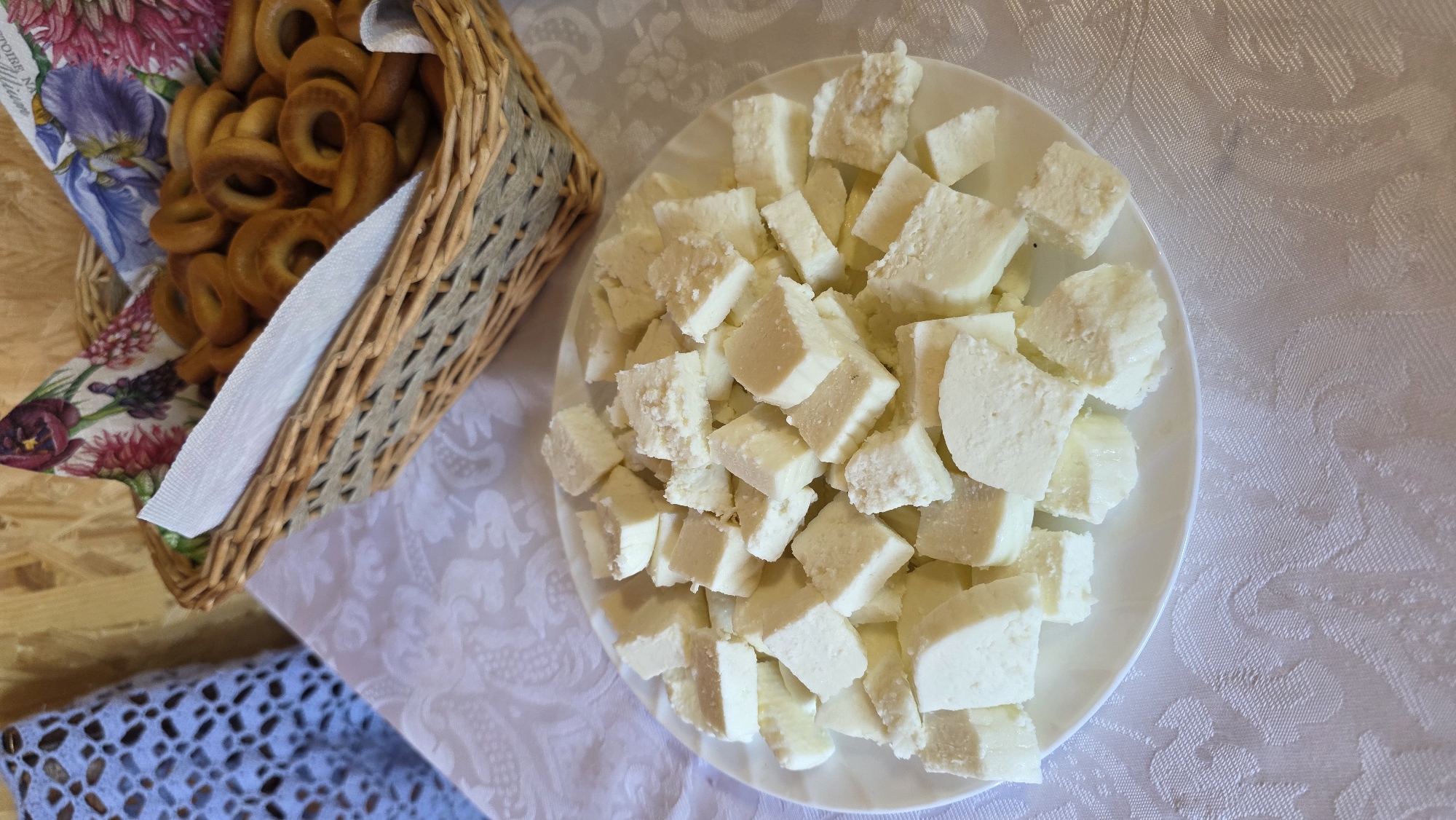 A plate of cubed white cheese sits on a white tablecloth patterned with embossed swirls. To the right of the plate, a woven basket filled with small, ring-shaped pastries is visible. The basket is lined with a paper napkin decorated with floral patterns. In the background, a light blue crocheted fabric item is partially visible on the left, and a wooden wall is behind the basket. The scene appears to be indoors, likely on a table. The location is specified as Alga, Russia.