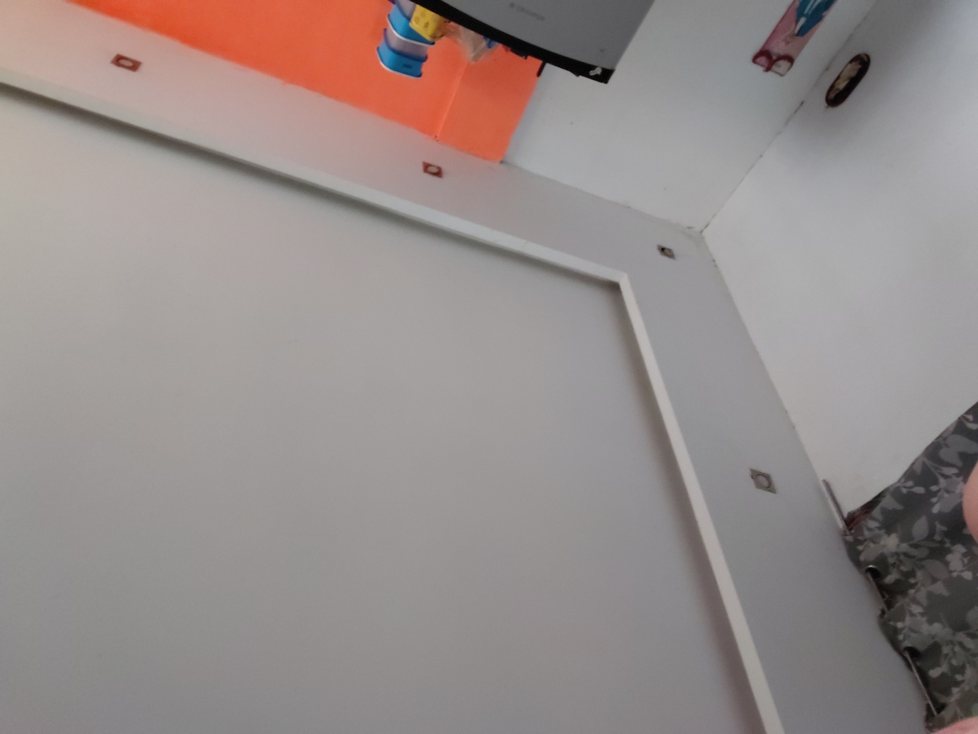 This image provides an upward, angled view inside a room in Santa Ana, Venezuela. A large, light-colored panel, possibly a wall or part of a piece of furniture, dominates the left and center, featuring a white border adorned with several small, square, dark brown fasteners or decorative elements.
Above this panel, a bright orange wall or structure is visible. Hanging from what appears to be a ceiling or the top of the orange fixture, there's a blue and yellow container, possibly a bottle, alongside a dark, rectangular electronic screen or small television that is mounted upside down.
To the right, a white wall meets the orange area. In the upper right corner, an oval-shaped, dark recessed fixture, likely a light or vent, is set into the white surface. Below it, a pink and blue fabric item or decoration is attached to the wall. The lower right corner of the image reveals a partial view of a person's shoulder or arm, covered by a grey and black patterned fabric, possibly clothing or a blanket. No visible text is clearly discernible on any of the objects. The scene suggests an informal indoor environment with an unusual arrangement for the electronic device.