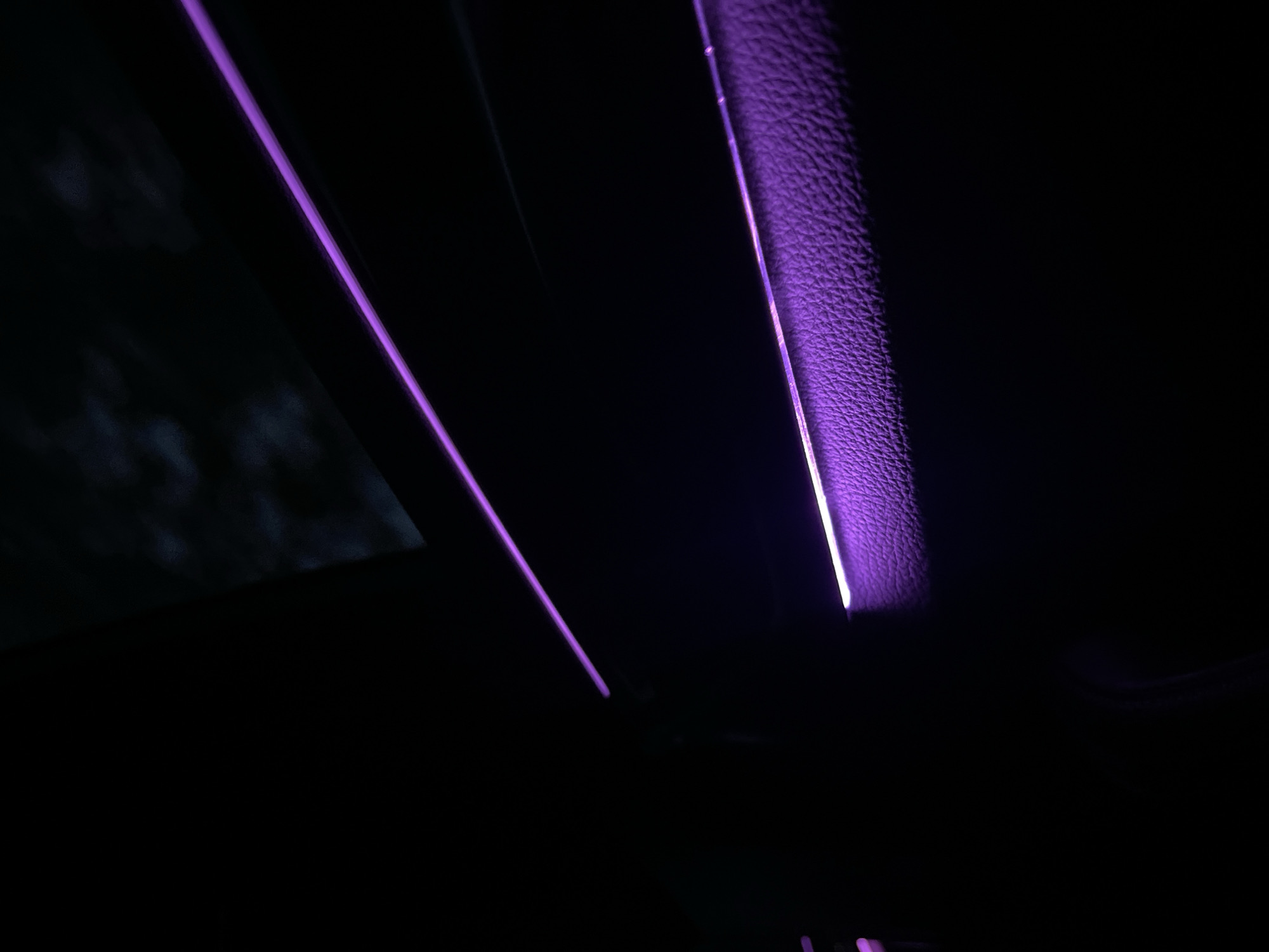 The image depicts a dark interior space, likely of a car, illuminated by purple lights. The texture of a material resembling leather is visible along the right side. The location is not discernible from the image.