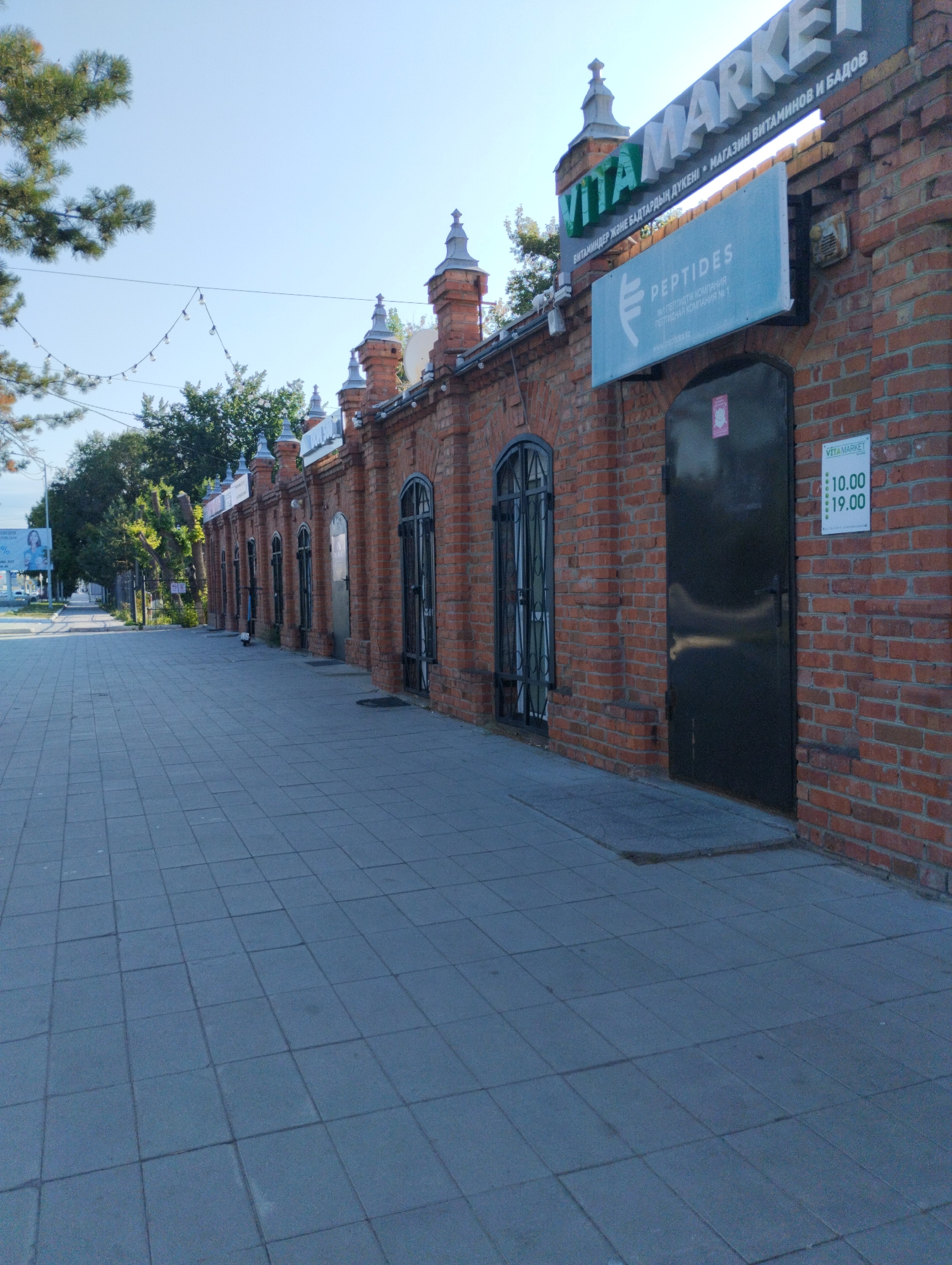 The image shows a street scene in Kostanay, Kazakhstan. A brick building with decorative architectural elements lines the right side of the frame. Above the entrance, a sign reads "VITA MARKET" in large, white letters, with smaller text underneath in what appears to be Kazakh. A lighter blue sign below that displays the word "PEPTIDES" and further text in Kazakh. A dark metal door is present on the right side of the building, and a sign with the VitaMarket logo and opening hours (10:00-19:00) is affixed to the brick wall nearby. The building has several arched windows with metal grates. A tiled sidewalk runs along the front of the building. To the left, trees and a street are visible, with a billboard in the distance. The sky is clear and blue. The overall impression is of a commercial street in a city.