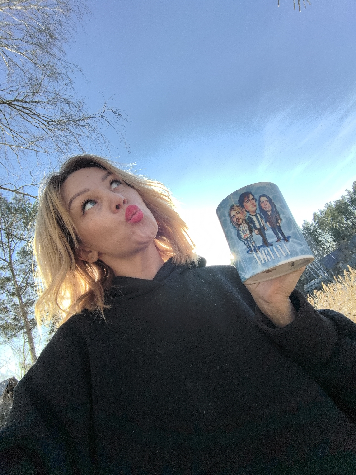 A woman is holding a mug with a caricature of the band Nirvana. The woman has blonde, shoulder-length hair and is wearing a black hoodie. She is pouting and looking up. The background includes trees, blue sky, and sunlight. The location appears to be outdoors. There is no direct evidence within the image to verify the location is in the city of Noviy, Russia.