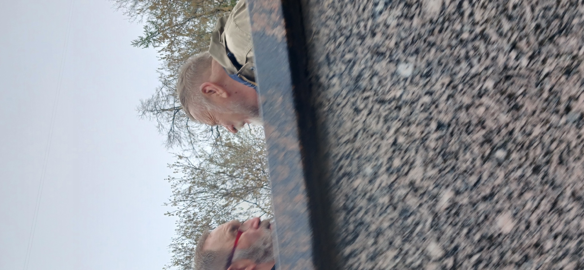 Two white males are standing near a granite structure. One man has short, gray hair and a beard and is wearing a tan jacket with a blue collar. The other man is wearing glasses with red frames and has a gray beard. Behind them are bare trees and a gray sky. The location is possibly a cemetery, Moscow, Russia.