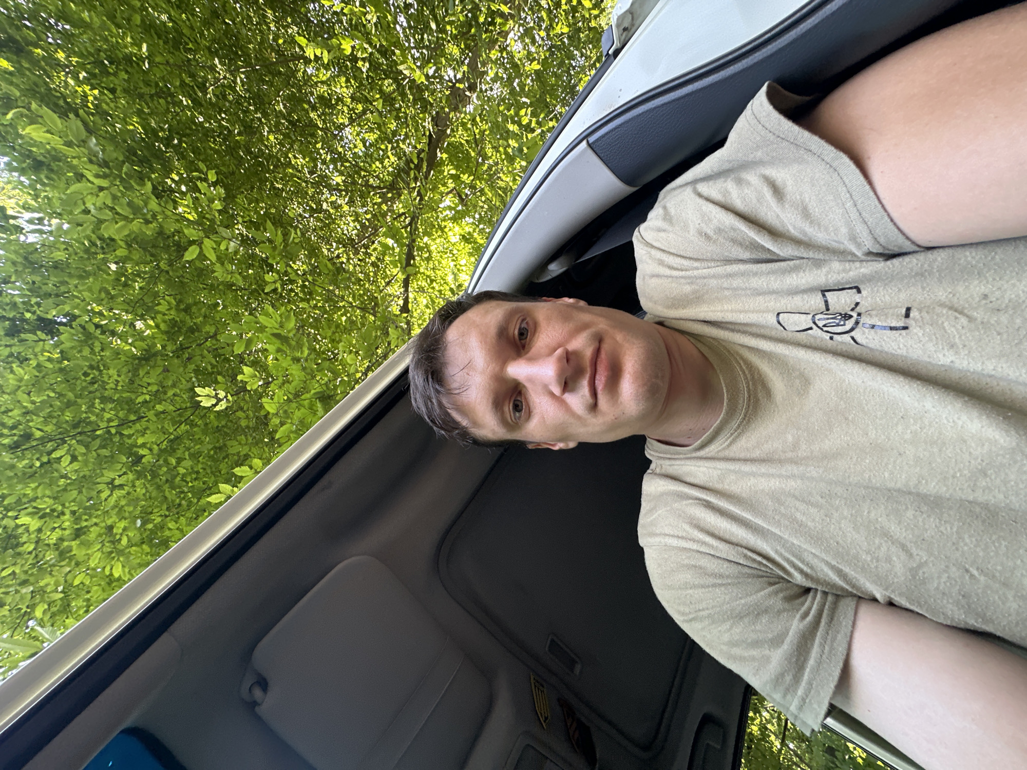 A man is sitting in the passenger seat of a car, looking directly at the camera. The car's sunroof is open, revealing a canopy of bright green leaves from overhead trees. The man is wearing a light tan t-shirt with a graphic on the left side of the chest. The interior of the car is visible, including a gray headliner and sun visor. The scene appears to be outdoors, likely in a wooded area.