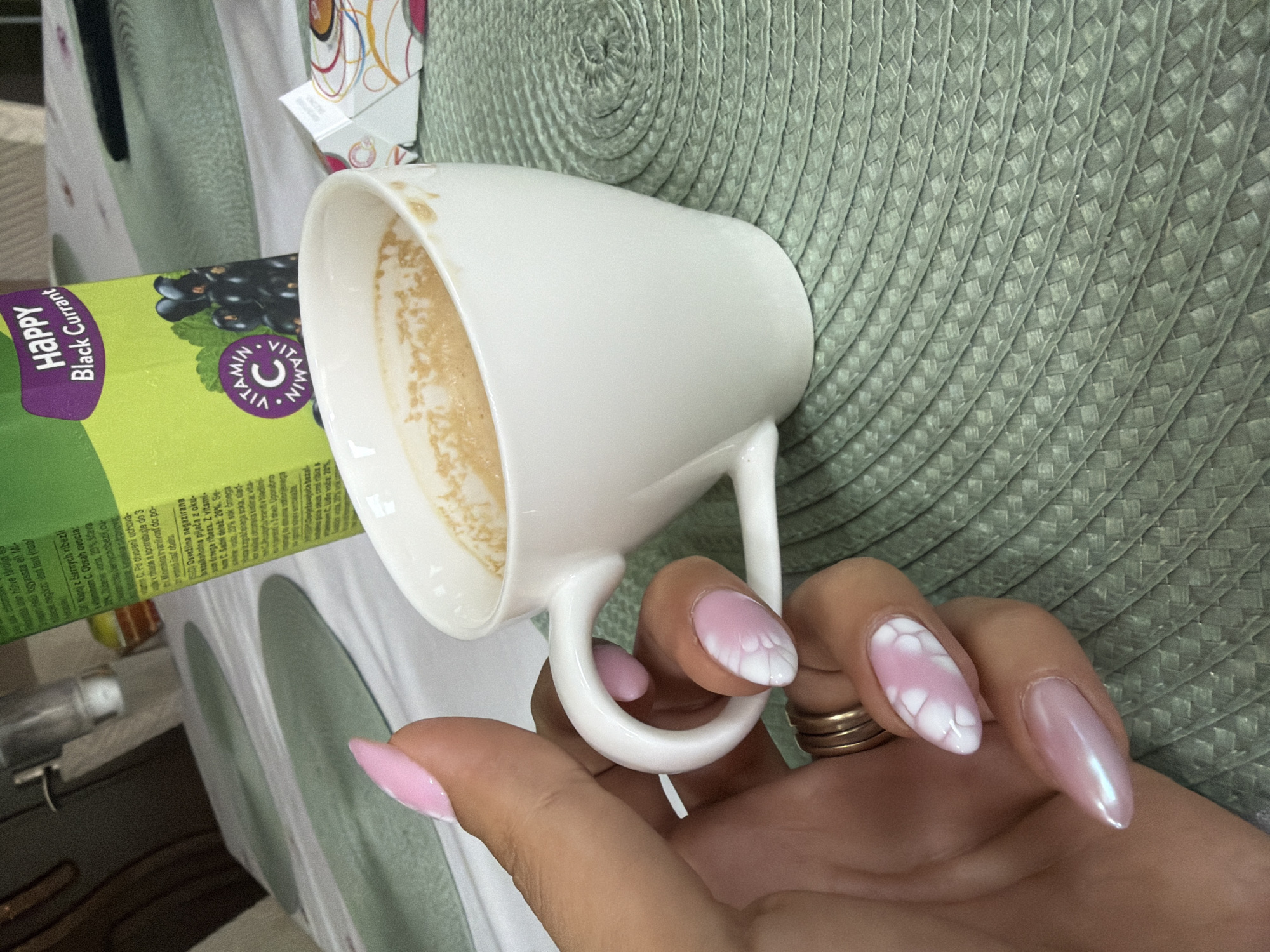 An angled, close-up photograph depicts a hand holding a white ceramic mug, which contains light brown liquid residue. The hand wears two gold-colored rings on one finger, and the nails are manicured with distinct designs: some are a solid glossy pink, while others feature a pink base with intricate white cloud-like or lace patterns.
To the left of the mug, a vibrant green carton of "Happy Black Currant" beverage is visible. The carton's label clearly states "Happy Black Currant" in purple text and displays an image of blackcurrants. A circular emblem highlights "VITAMIN C." Text on the packaging, including phrases like "Po otvorení uchováva" (After opening store...) in Slovak/Czech and "Osvežilna negazirana brezalkoholna pijača z okusom črnega ribeza. Z vitaminom C" (Refreshing non-carbonated non-alcoholic beverage with blackcurrant flavor. With Vitamin C.) in Slovenian, indicates the product's likely market.
The mug and the drink carton are placed on a textured, light green, circular woven placemat, which forms the primary background. The scene is set indoors, suggesting a casual home environment like a kitchen or dining area in Plovdiv, Bulgaria. The image captures a quiet moment, focusing on the details of the hand, mug, and beverage carton.