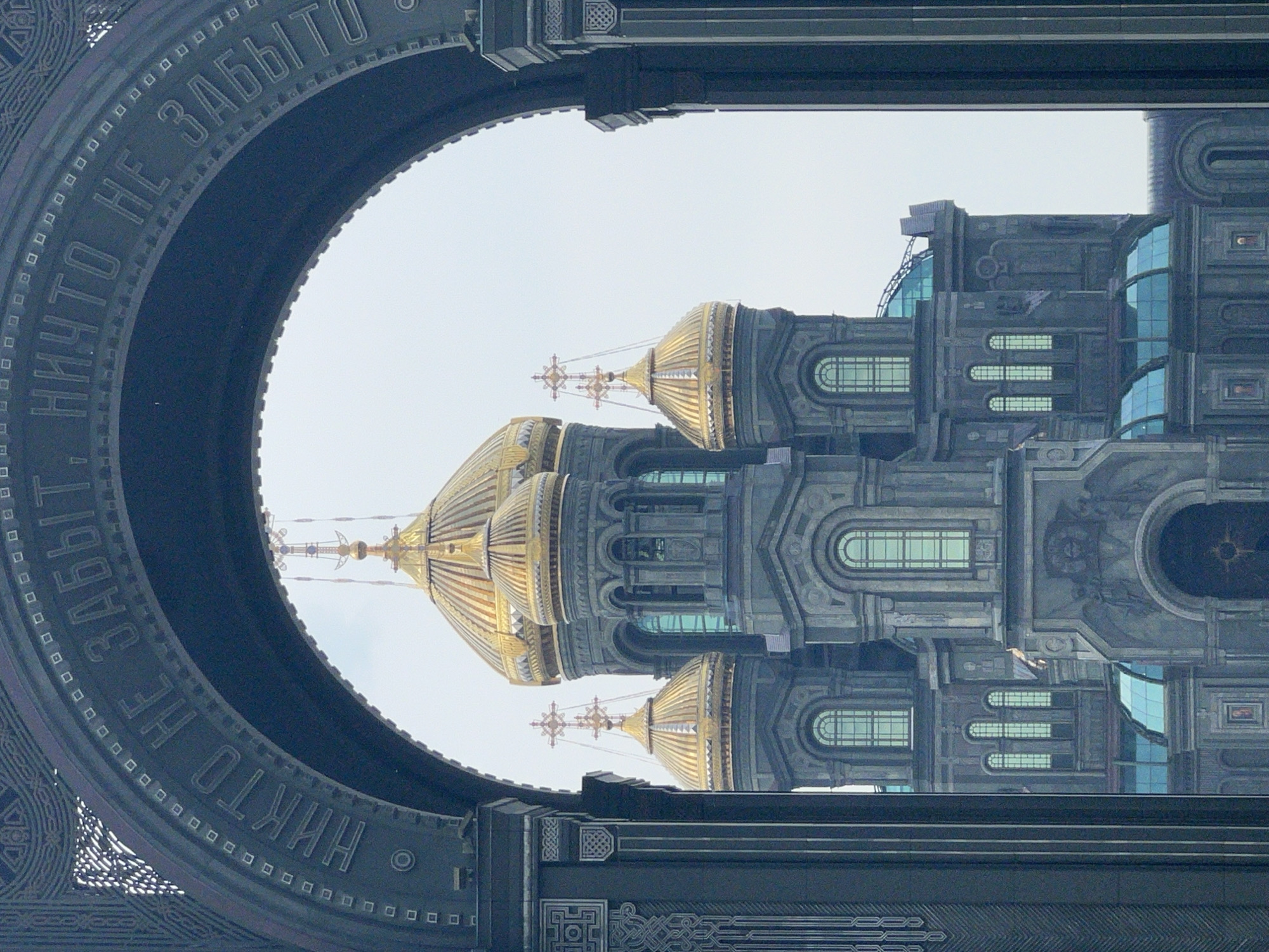The image captures a view of a grand, gray, multi-domed cathedral with golden roofs, seen through a dark, arched architectural element. The cathedral has several layers of windows, some arched and some rectangular, and ornate detailing. Inscriptions are visible on the dark arch, reading in Russian "НИКТО НЕ ЗАБЫТ, НИЧТО НЕ ЗАБЫТО," which translates to "No one is forgotten, nothing is forgotten." The sky is overcast and light. The architectural style of the cathedral suggests it is a significant religious or historical monument.