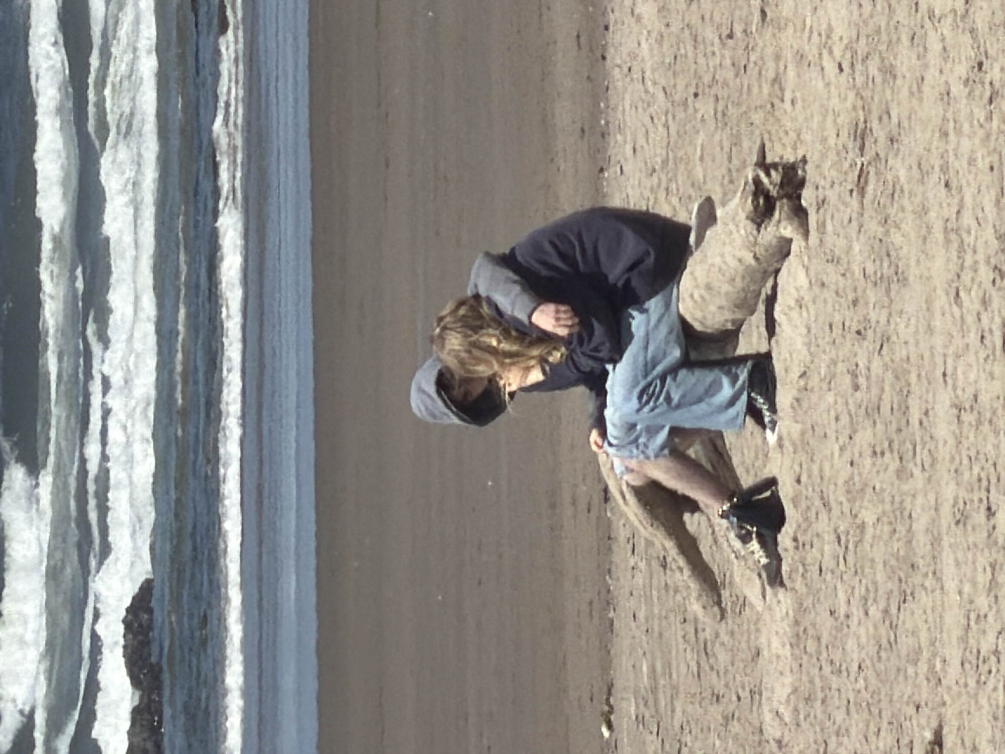 Two individuals, one with blonde hair and wearing a dark hoodie and light blue jeans, the other with a hood over their head, are embracing while sitting on a piece of driftwood on a sandy beach. The person with blonde hair is positioned in front, with their head tilted back. The other individual's arm is around their shoulders. The ocean with waves is visible in the background. The scene appears to be on a beach in Cannon Beach, Oregon, United States of America.