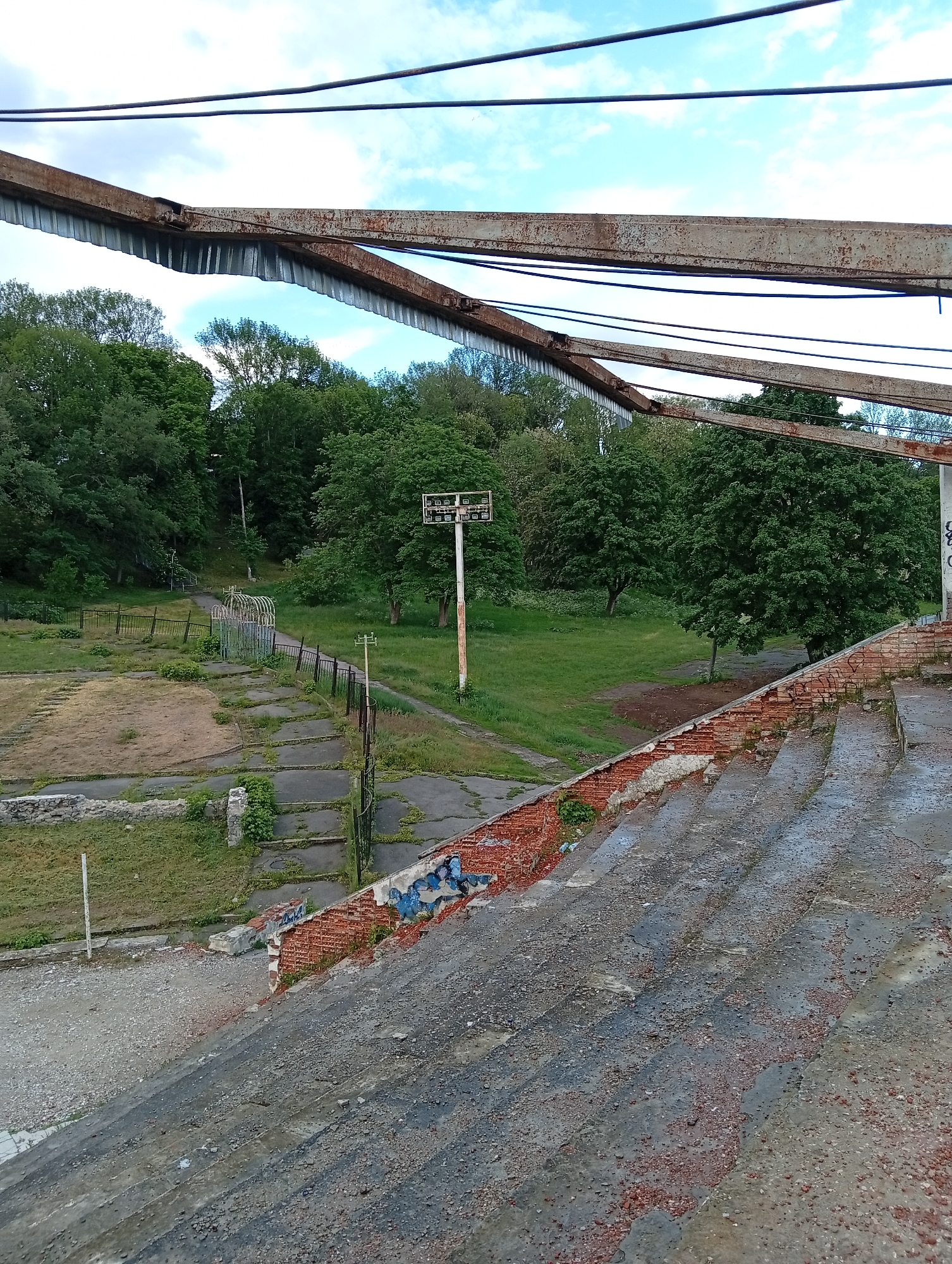 The image depicts a dilapidated outdoor stadium in Poltava, Ukraine. The seating area is constructed of crumbling concrete and brick, with significant deterioration and discoloration. Vegetation grows in the cracks and crevices of the structure. An old, rusted metal frame structure overhangs the stands. Beyond the stands, a grassy field is visible, along with an old metal fence and a tall pole with lights. Trees surround the stadium, and the sky is visible in the background with some clouds. There are no people visible.