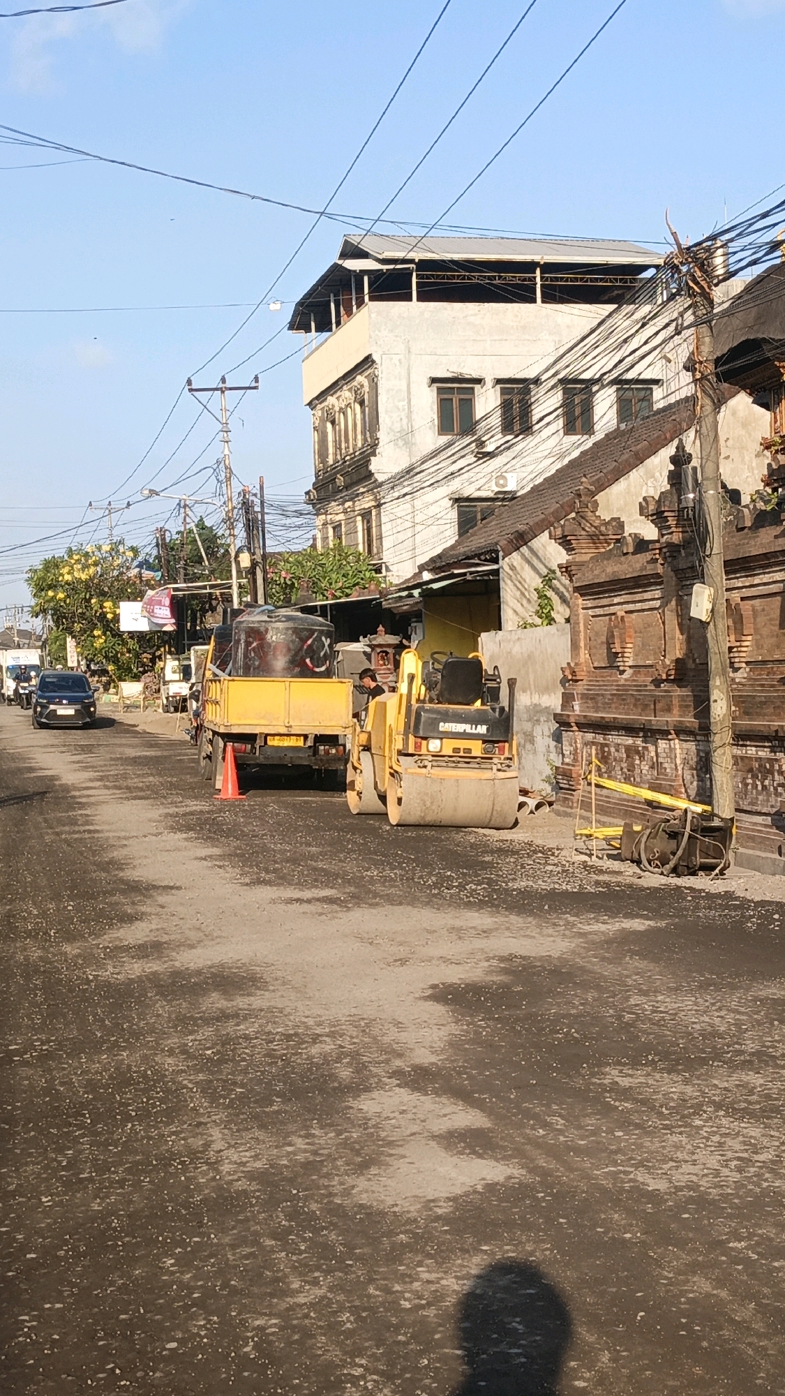 The image depicts a street scene in Denpasar, Indonesia, likely during a road construction or repair project. A yellow Caterpillar steamroller is positioned on the asphalt, with a yellow dump truck behind it. The dump truck contains a large black water tank with graffiti. An orange traffic cone is placed near the truck. The road surface appears to be freshly laid or disturbed asphalt, with loose gravel visible. In the background, a multi-story building with a light-colored facade and a dark roof stands prominently. There are numerous electrical wires overhead, a common sight in urban Indonesian settings. To the right, a traditional Balinese-style brick wall and a wooden pole with electrical fixtures are visible. The sky is clear and blue, suggesting it is daytime. A shadow of a person is cast on the road in the foreground, indicating the photographer's presence. A few vehicles are visible in the distance, suggesting ongoing traffic.