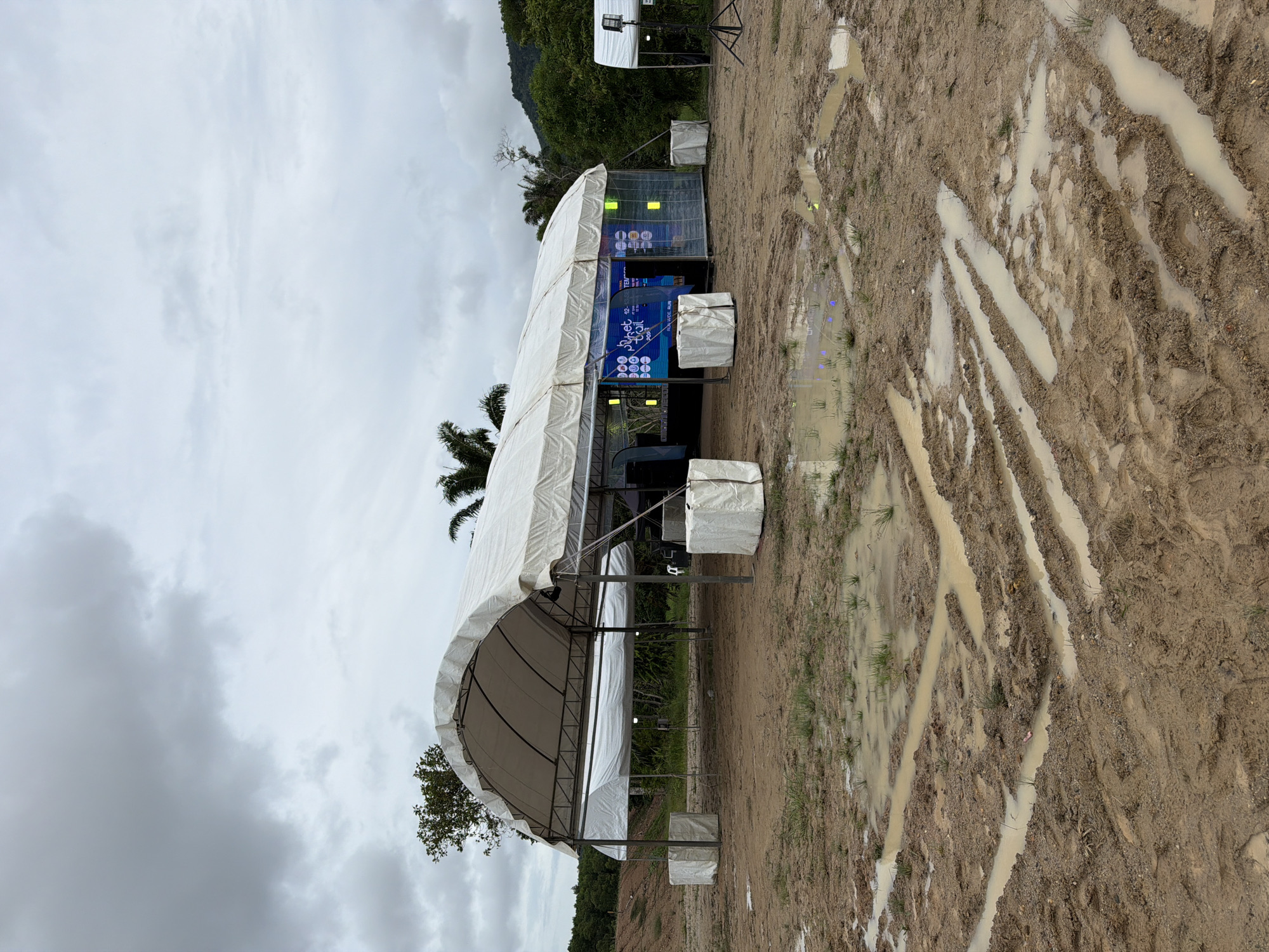 An outdoor event setup is visible under a cloudy sky in Si Sunthon, Thailand. The scene is characterized by a large, white, arched tent with a metal frame. The ground is muddy with several puddles, indicating recent rainfall. Tire tracks are visible in the mud, suggesting vehicle activity.
Inside the tent, a stage area is set up with a large screen displaying graphics. The screen appears to be part of a promotional display, with text and logos visible, possibly related to an event like a run or trail race, as suggested by phrases like "Phuket Trail" and "Run Wide Run." There are also lights and other equipment associated with an event setup.
Several white, cube-shaped structures covered in fabric are placed around the tent, likely serving as weights or ballast for the tent structure. Two smaller tents are also visible, one behind the main tent and another to the right, with a tripod-mounted light. The surrounding environment includes trees, vegetation, and a gentle slope of land to the left. The overall impression is of an event venue that is either being prepared, is in use, or has recently concluded, with the muddy conditions suggesting it's an outdoor location susceptible to weather. There are no people visible in the image.