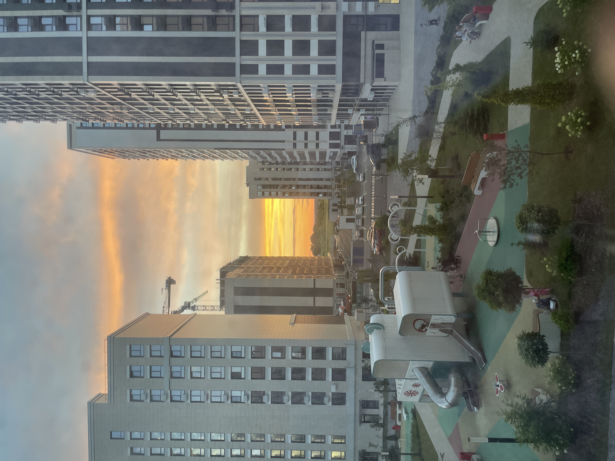 This media file depicts an aerial view of a residential complex in Stavropol, Russia, during sunset. The scene is dominated by modern, multi-story apartment buildings with numerous windows reflecting the warm hues of the sky. In the foreground, a playground with slides, swings, and benches is visible, suggesting a family-friendly environment. Several people are present on the playground, enjoying the late afternoon. The sky is ablaze with vibrant orange and yellow tones, characteristic of a sunset, casting a warm glow over the urban landscape. Construction cranes are also visible in the distance, indicating ongoing development in the area. The overall atmosphere appears peaceful and serene, with the beauty of nature contrasting with the man-made structures. There is no visible text in the image.