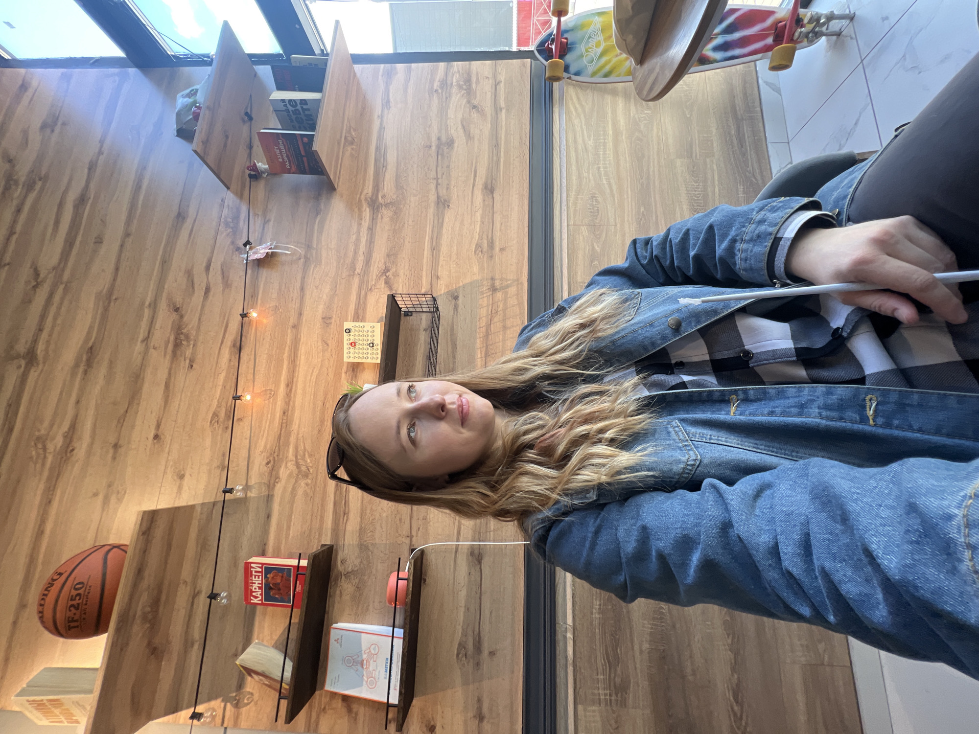 A woman with long blonde hair and a denim jacket sits in what appears to be a cafe or bookstore in Mytishchi, Russia. Behind her are wooden paneled walls with shelves displaying books and objects like a basketball and a small sculpture. A skateboard hangs near the ceiling. The woman is holding a white object, possibly a pen or cigarette. The location is identified by the Cyrillic text on the books on the shelves, and the Russian appearance of the woman.