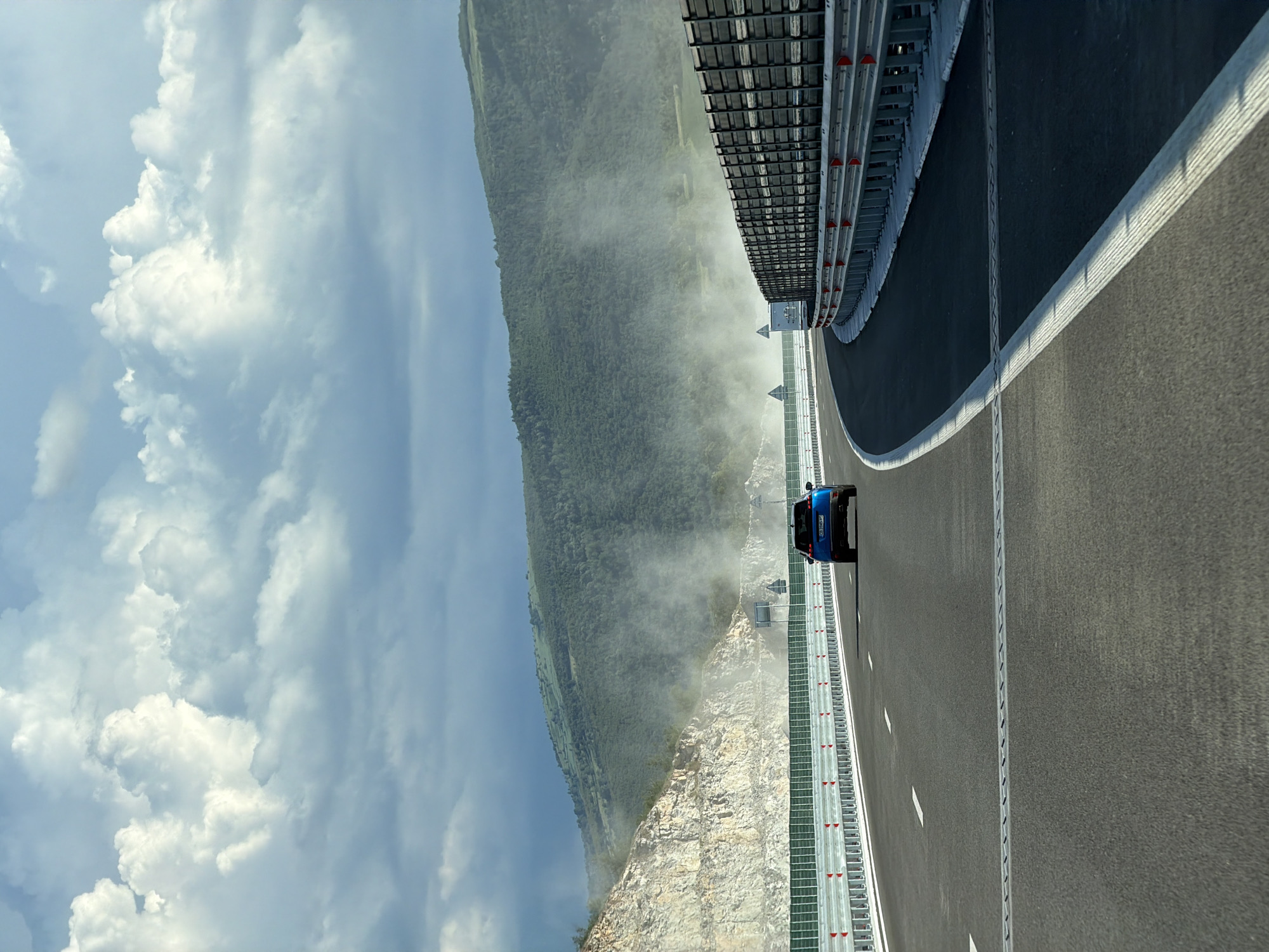 The image depicts a highway, likely a new construction or under development, curving through a mountainous landscape. A blue car is seen driving on the highway, facing away from the camera. The road is asphalt with clear lane markings and safety barriers on either side. The left side of the highway features a steep, rocky incline with visible layers of stone, suggesting excavation or a cut through the mountainside. The right side of the highway is bordered by a dark, tall sound barrier. In the background, lush green hills are covered in dense forests. A hazy mist or dust is visible in the air, particularly near the rocky incline and between the highway and the hills, which could indicate ongoing construction activity or natural conditions. The sky is partly cloudy with dramatic, large white clouds against a lighter blue sky, suggesting dynamic weather conditions. The scene appears to be set on the outskirts of Peshtera, Bulgaria, given the context of recent highway development in the region.
