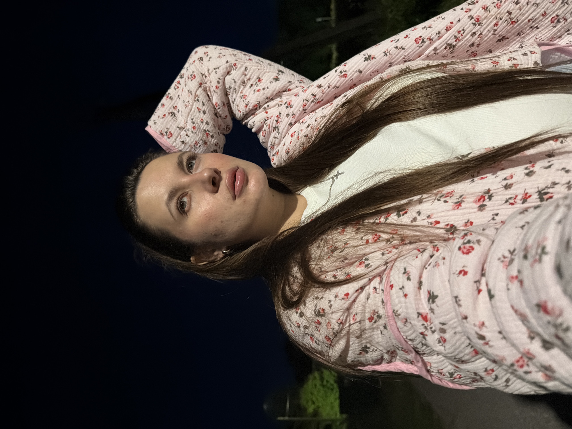 A young woman with long, dark brown hair is shown outdoors at night. She is wearing a pink, floral-patterned quilted jacket over a white t-shirt. Her right arm is bent, and her hand is resting on the back of her head. She is looking up and to her right. The background is dark, with a few faint lights visible in the distance, suggesting an urban or suburban environment. The specific location in Krasny Mayak, Russia, is not discernible from the image itself, but the scene depicts a person in a casual pose in an outdoor setting at night.