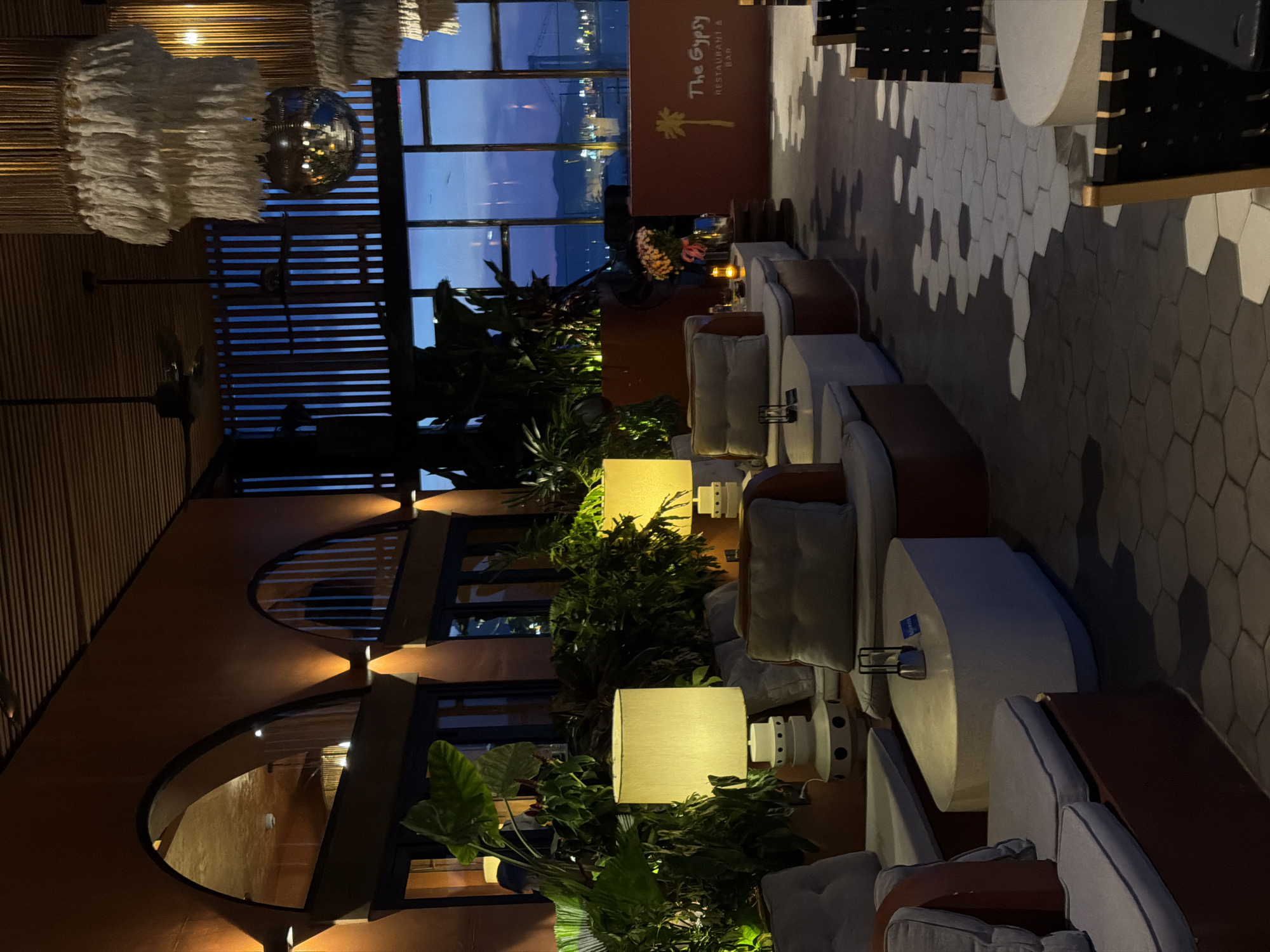 The image depicts the interior of a restaurant and bar called "The Gypsy," located in Đà Nẵng, Vietnam. The scene is dimly lit with warm ambient lighting, creating a relaxed and sophisticated atmosphere.
In the foreground, there are plush, low-slung modular sofas arranged around oval-shaped coffee tables. These seating areas are upholstered in light gray fabric with dark red accents on the sofa bases. Two large table lamps with cream-colored shades are placed near the sofas, providing localized illumination. The floor is covered with dark gray concrete tiles featuring a hexagonal pattern that transitions to lighter gray and white tiles in certain areas, creating a dynamic visual effect.
Behind the seating area, large windows offer a view of the city at dusk or dawn, with lights from buildings and structures visible across a body of water. The sky displays hues of deep blue and purple.
The restaurant's interior features a ceiling with dark wooden slats and exposed beams. A disco ball hangs from the ceiling, reflecting light and adding a touch of retro flair. Several large ceiling fans are also visible. Decorative pendant lights with white macrame fringes hang above the seating areas.
The walls are painted in a warm, terracotta orange color, and architectural elements like arched windows are present. Lush green plants, including large-leafed varieties, are strategically placed throughout the space, adding a natural and tropical feel.
On the right side of the image, a reception desk or bar area is visible. It is a bright orange structure with the restaurant's name, "The Gypsy," and "RESTAURANT & BAR" printed on it in a stylized font, accompanied by a palm tree graphic. A bouquet of colorful flowers in a vase sits on a small table next to the desk.
A few chairs with black woven seats and wooden frames are placed in the middle ground, suggesting additional seating arrangements. The overall impression is a stylish and comfortable venue, likely popular for evening dining and drinks. No people are clearly identifiable in the image, though a blurred figure might be present in the background.
