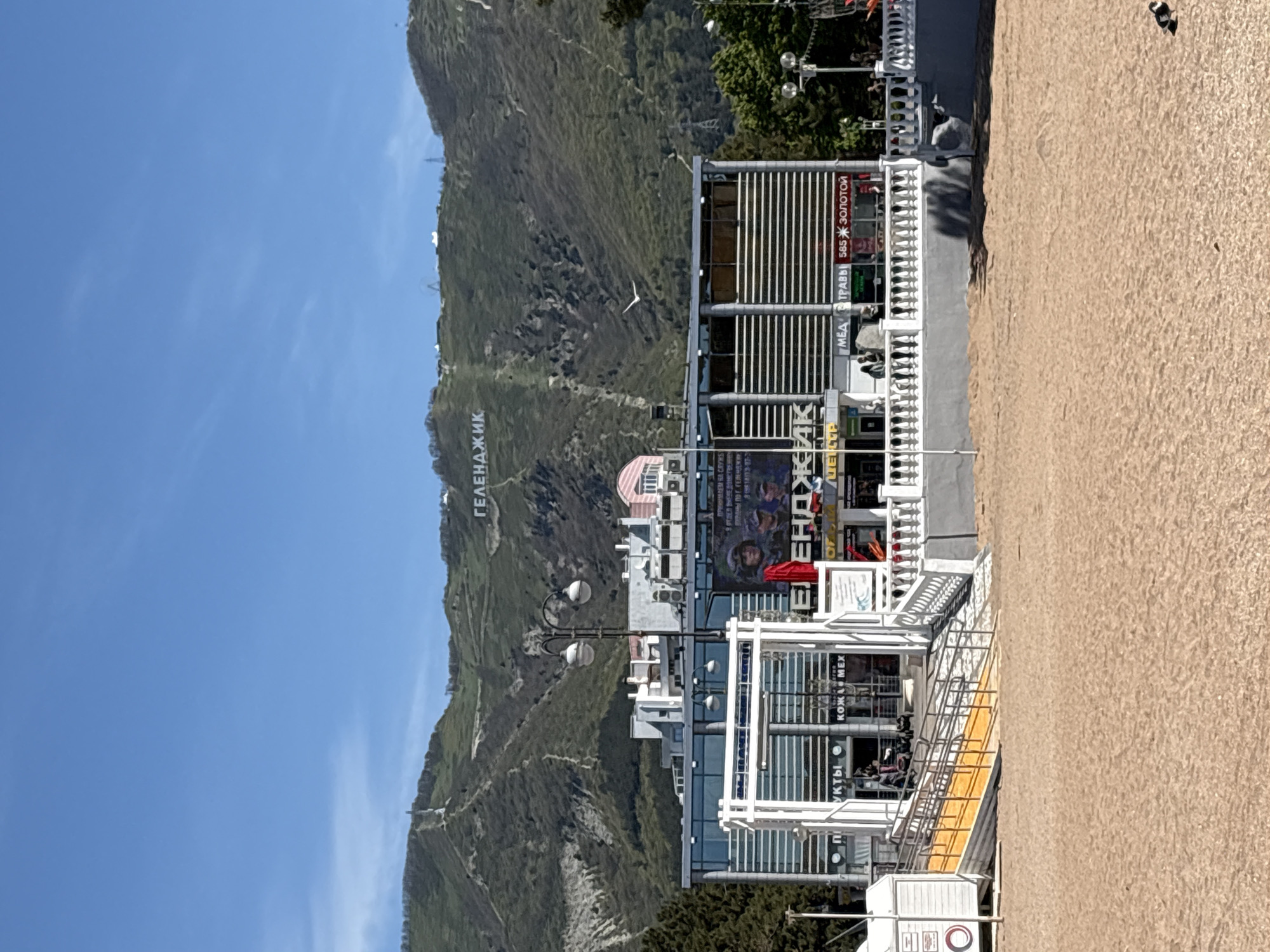 This image depicts a sunny daytime scene in Gelendzhik, Russia, featuring a bustling urban area set against a large, verdant mountain.
In the background, a prominent mountain or hill, thickly covered with green vegetation, rises under a clear blue sky with a few faint wisps of clouds. Large white letters spelling "ГЕЛЕНДЖИК" (Gelendzhik) are visible on the mountainside, identifying the location. A lone white bird is seen flying against the mountain.
In the mid-ground, a series of modern buildings with large windows and white facades line the base of the mountain. These structures appear to house commercial establishments, with visible signs such as "КОЖА МЕХ" (Leather Fur), "МЕД ТРАВЫ" (Honey Herbs), and "585 ЗОЛОТОЙ" (585 Gold). Other partial text includes "ЕНДЖИК," "УКТЫ," and phrases like "ПРИНИМАЕМ НА С..." and "ВПИРИНЫ ПО Г. ГЕЛЕНДЖИК," indicating various services or products. White railings and ornate lampposts decorate the area around the buildings.
The foreground consists of a wide, sandy area, likely a beach or promenade, stretching out prominently. A few indistinct figures of people are visible near the buildings and on the sandy path. The bright sunlight creates clear shadows, suggesting it is midday or early afternoon. The overall impression is that of a lively coastal resort town on a pleasant day.