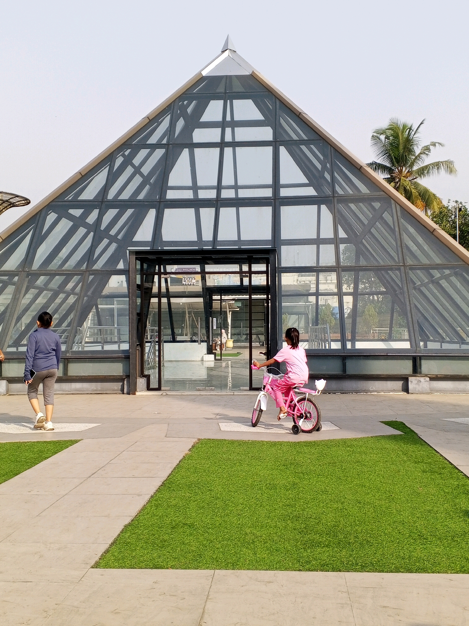 In Serang, Indonesia, a pyramid-shaped glass building with a steel frame stands under a clear sky. A young girl, wearing a pink t-shirt and pants, rides a pink bicycle with training wheels on a paved area in front of the building. Another person, dressed in a blue long-sleeved shirt and gray pants, walks away from the building, looking at a phone. Green grass patches are interspersed with the paving stones. A palm tree is visible to the right of the pyramid.