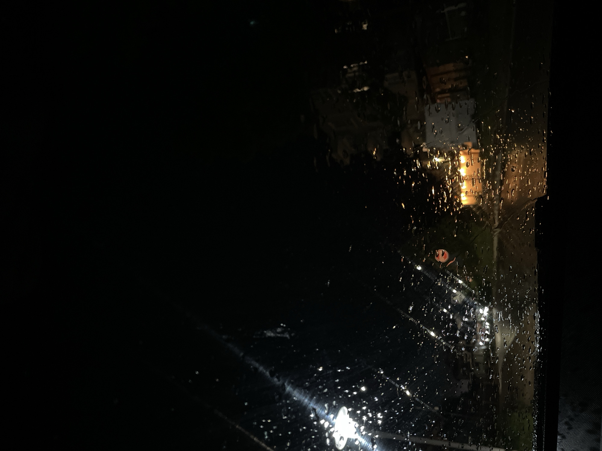 The image depicts a view through a rain-streaked window at night. Outside, blurred lights suggest a street scene. There is a glimpse of a building or structure with a warm, illuminated interior. The darkness obscures the finer details of the surroundings. It is not possible to ascertain the location as Byala, Bulgaria from the image alone.