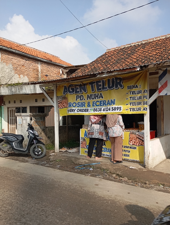 An outdoor scene in the city of Situwangi, Indonesia, depicts an egg vendor stall situated in front of a building with a red-tiled roof. The stall features a prominent yellow banner displaying "AGEN TELUR" (Egg Agent), "PD. NUHA", "GROSIR & ECERAN" (Wholesale & Retail), a delivery order number, and a list of available egg types including chicken, quail, and duck eggs. Multiple crates of brown eggs are displayed on a counter within the stall. Two individuals, likely women, are positioned with their backs towards the viewer, facing the egg display, one wearing a patterned top and dark pants, the other a light brown hijab and light-colored trousers. To the left of the stall, a dark gray scooter is parked on the unpaved ground adjacent to an asphalt road. The background shows a second building with exposed brickwork and a red-tiled roof. The sky is clear blue with scattered white clouds, and overhead power lines are visible. Empty egg cartons are scattered on the ground near the stall, and general debris is present at the roadside. A vertical red and white striped pole, possibly a barber shop sign, is partially visible on the far right.