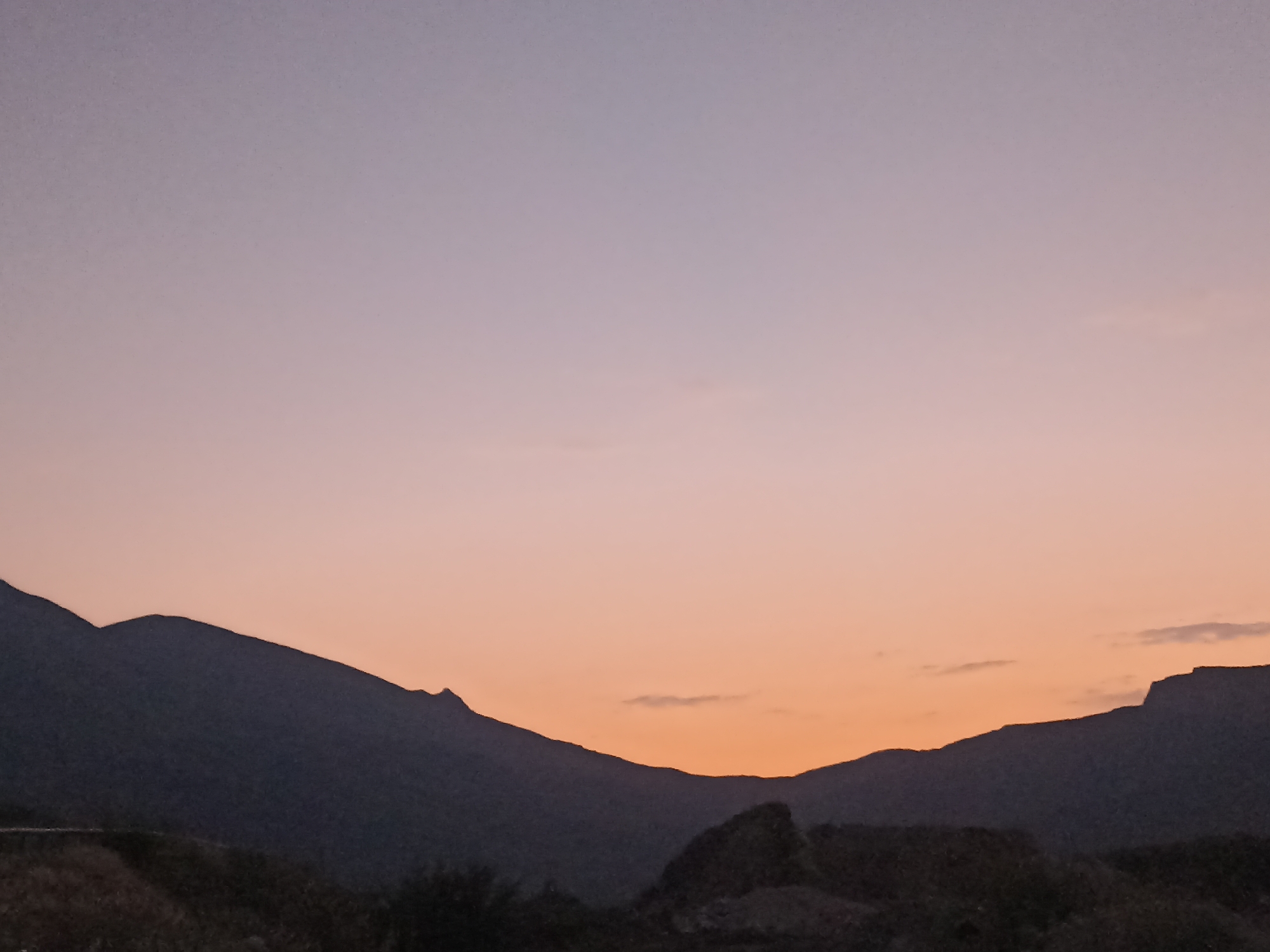 The image depicts a silhouette of mountains against a twilight sky. The sky transitions from a pale purple at the top to a warm orange hue near the horizon, suggesting either dawn or dusk. The mountains are dark and their jagged outlines are clearly defined against the lighter sky. In the foreground, the ground appears barren and rocky, with some dark vegetation visible. There are no people or animals present in the image. The scene is serene and natural, with a focus on the interplay of light and shadow in a mountainous landscape. This image captures a view of the natural environment surrounding the city of Kuzun, Azerbaijan.